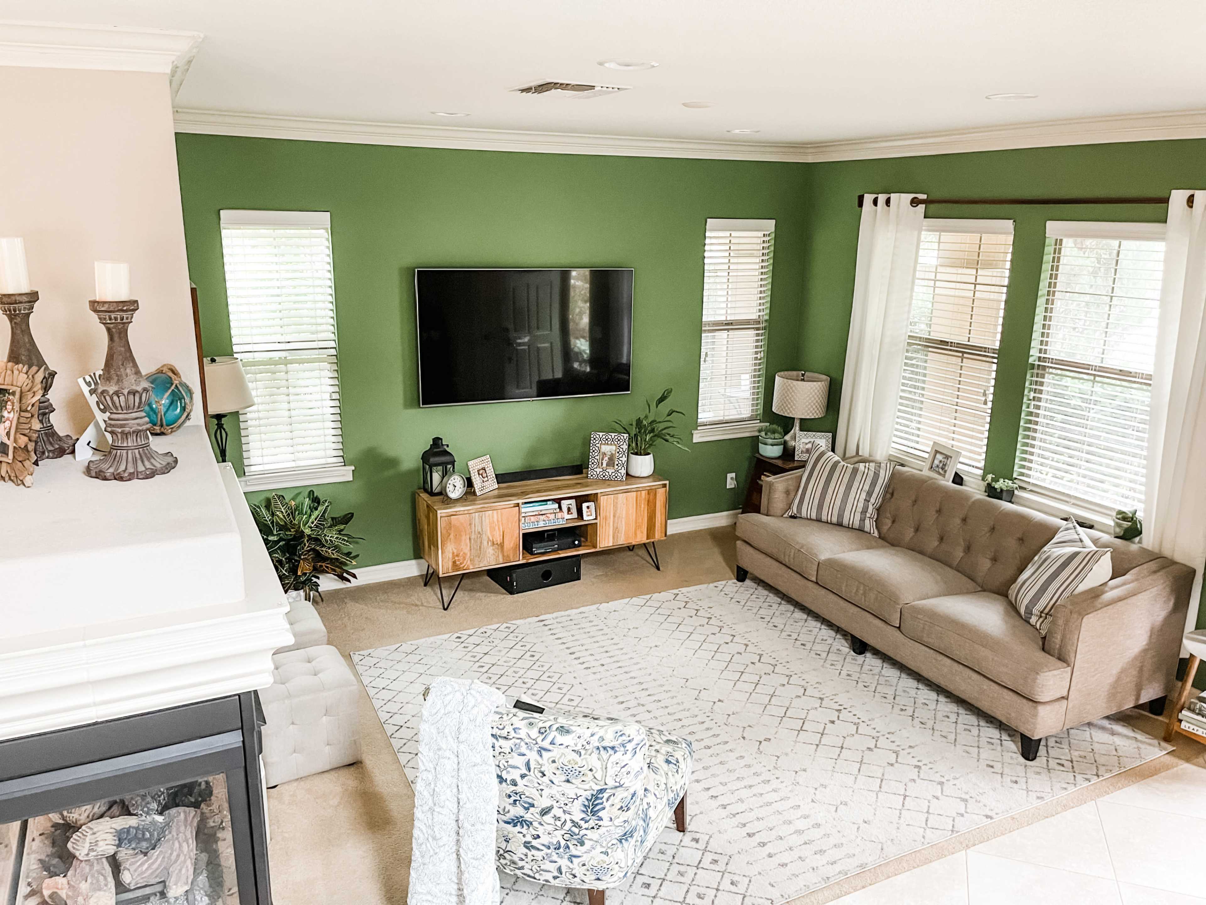 A living room features a green accent wall, a large TV mounted above a wooden sideboard, a beige sofa, and a patterned area rug, with large windows allowing natural light to enter.