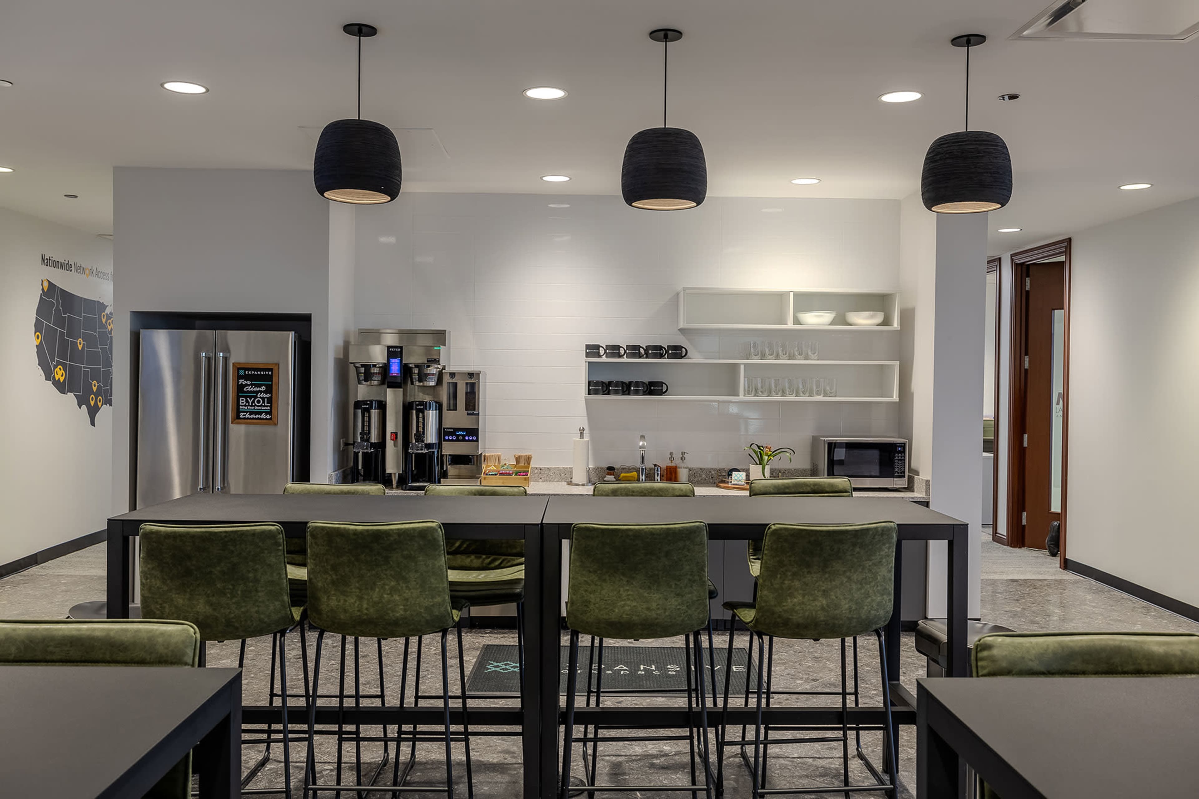 The image shows a modern kitchen area with high tables, green bar stools, and stainless steel appliances, featuring a coffee station and a wall-mounted map.