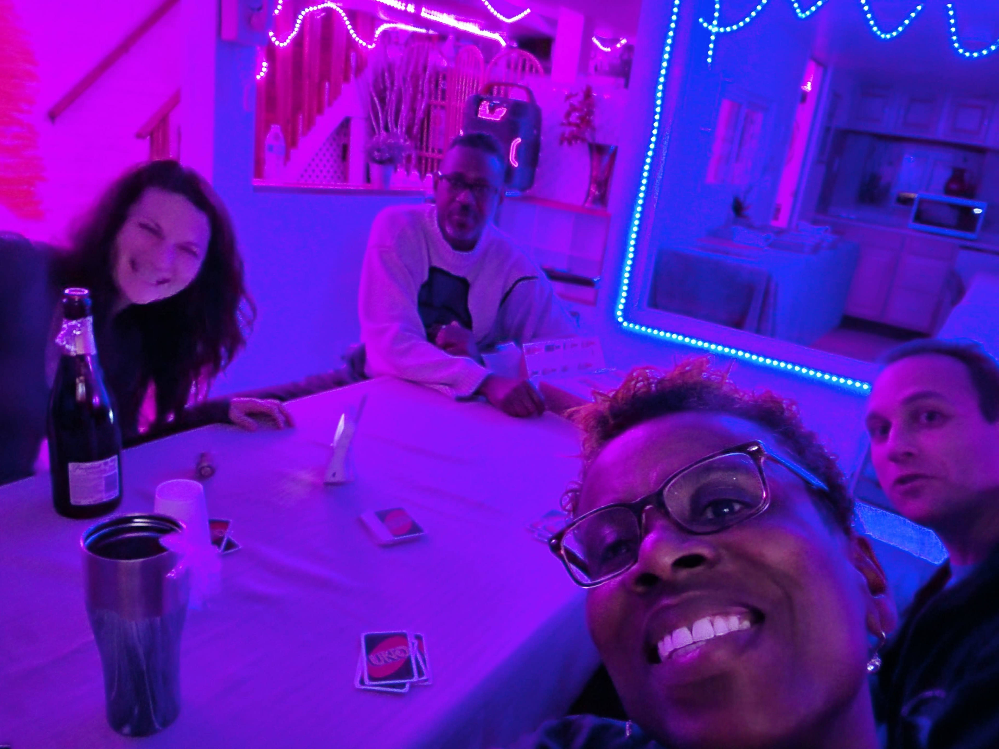 A group of four people gathers around a table illuminated by colored neon lights in a room.