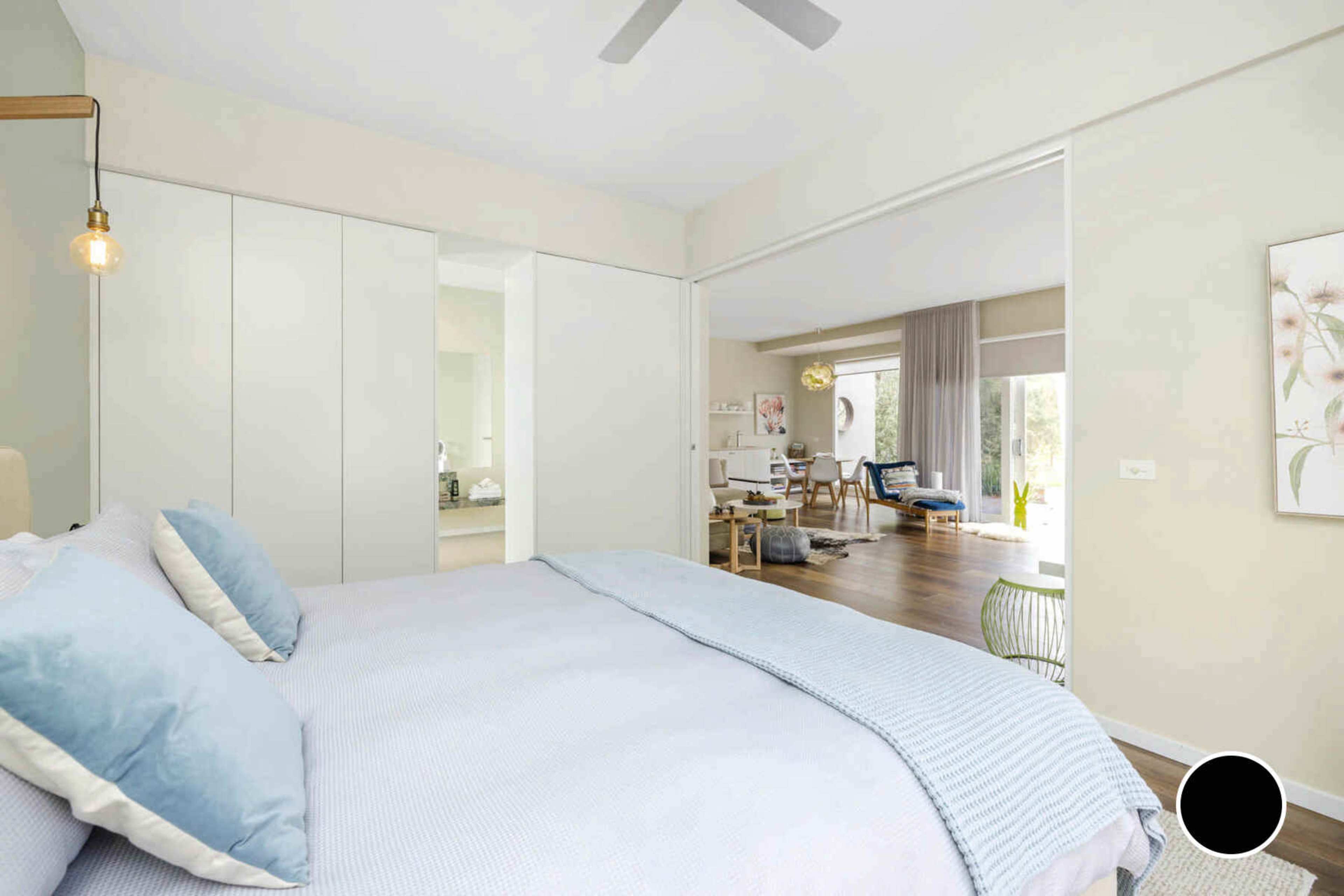 A well-lit bedroom features a bed with light blue linens, a modern closet, and a view into a living area through an open sliding door.