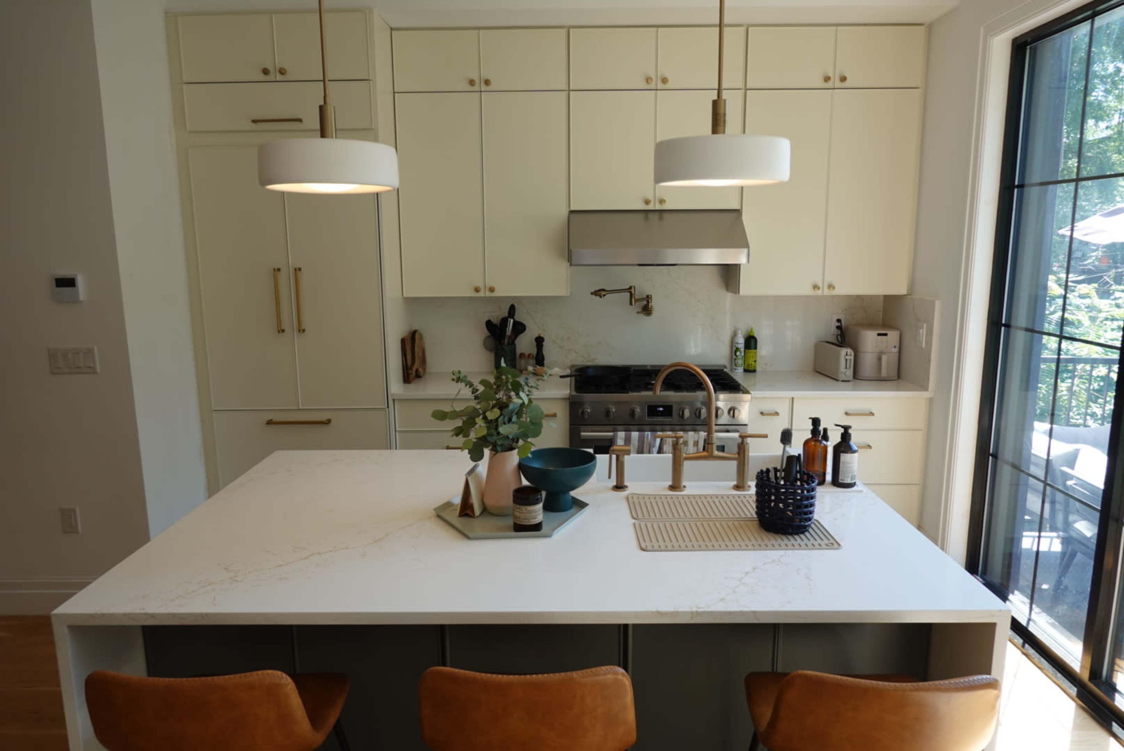 The image shows a modern kitchen with white cabinetry, a central island, a stainless steel stove, and large windows overlooking greenery.