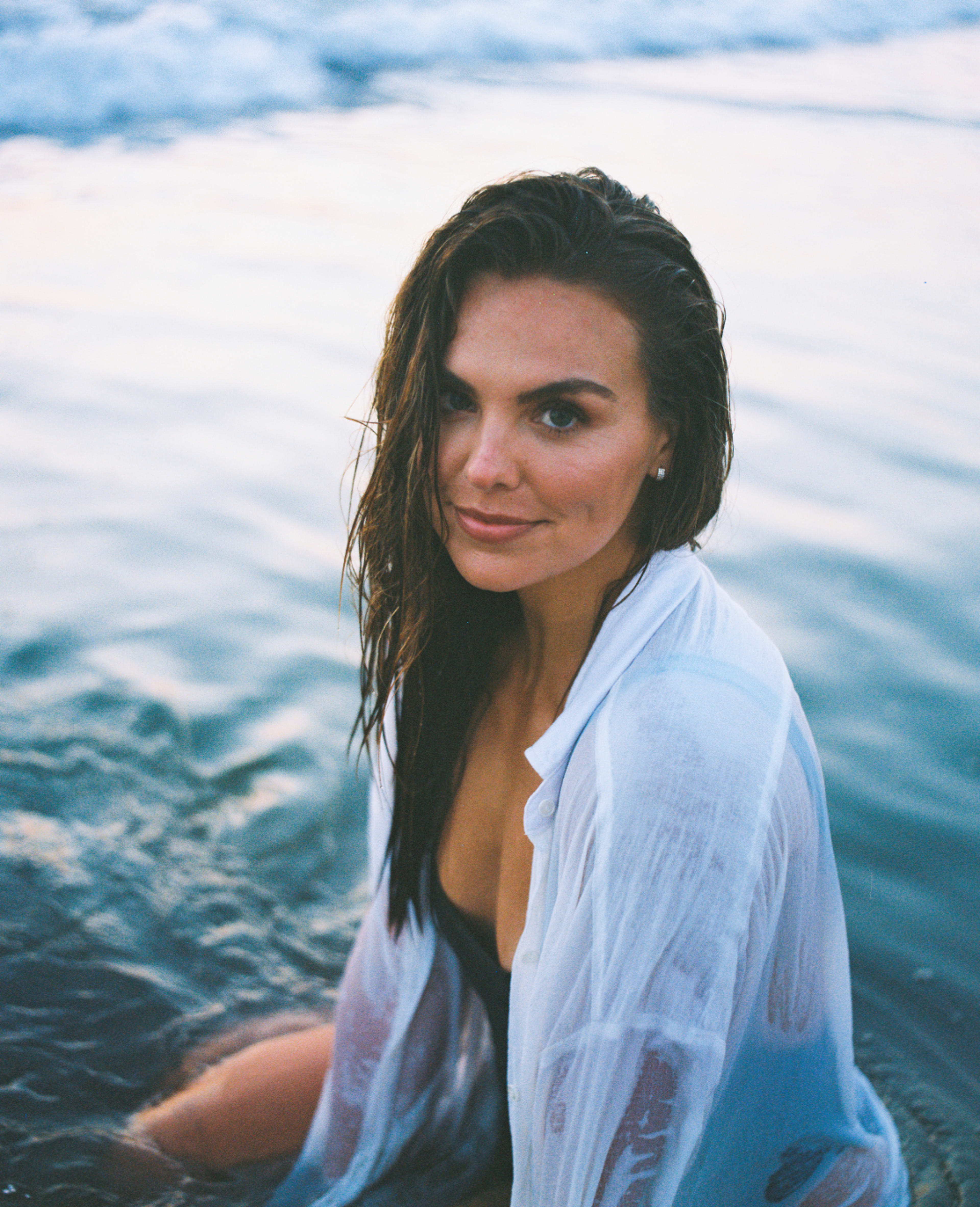 A woman sits in shallow water at the beach, wearing a wet white shirt and a swimsuit, with ocean waves in the background.