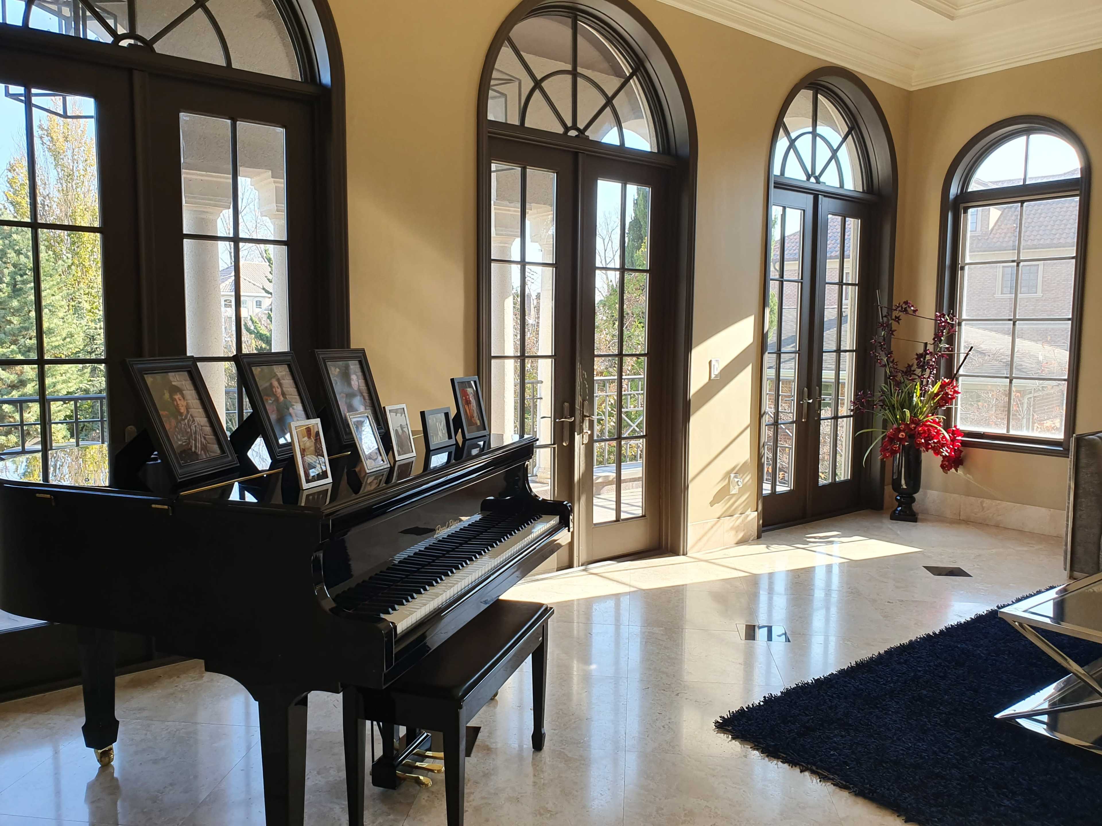 A black piano sits beside a wall adorned with photo frames, illuminated by natural light streaming through large arched windows in a spacious room.