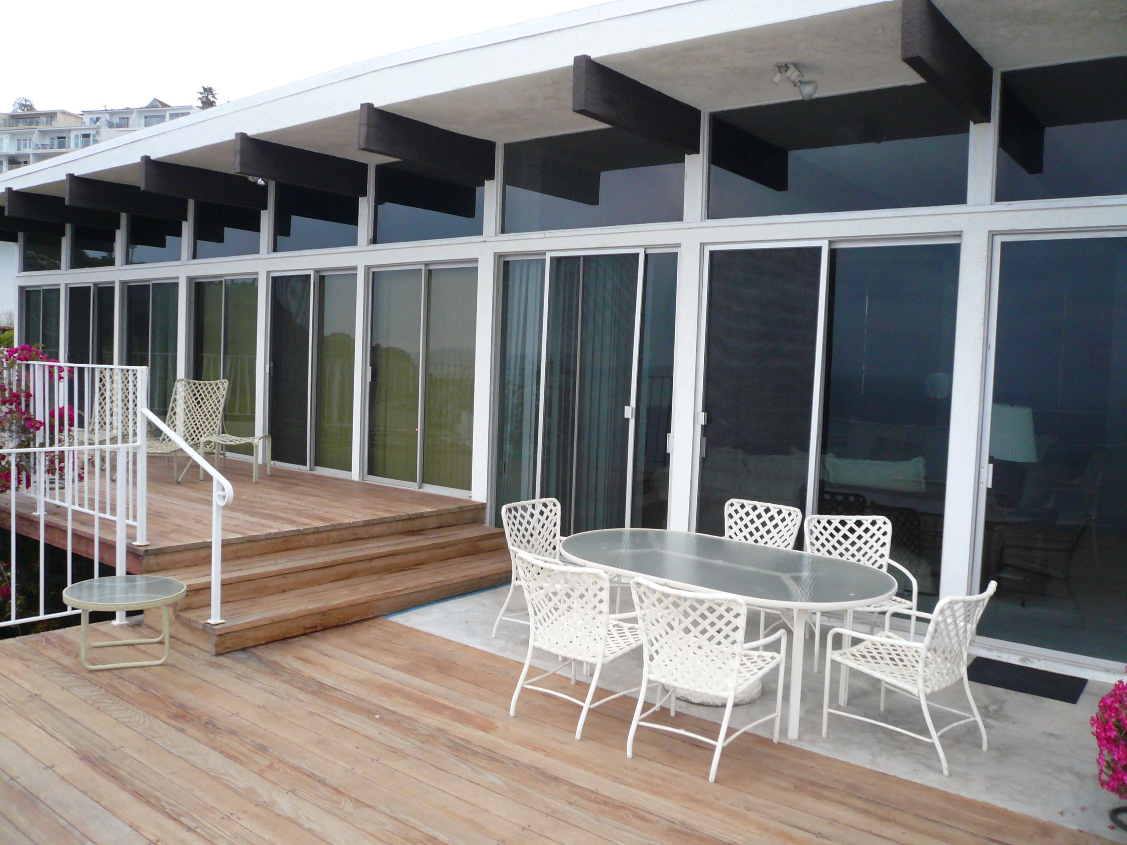 A patio with a glass table and white chairs is situated in front of a row of large windows at a modern building.