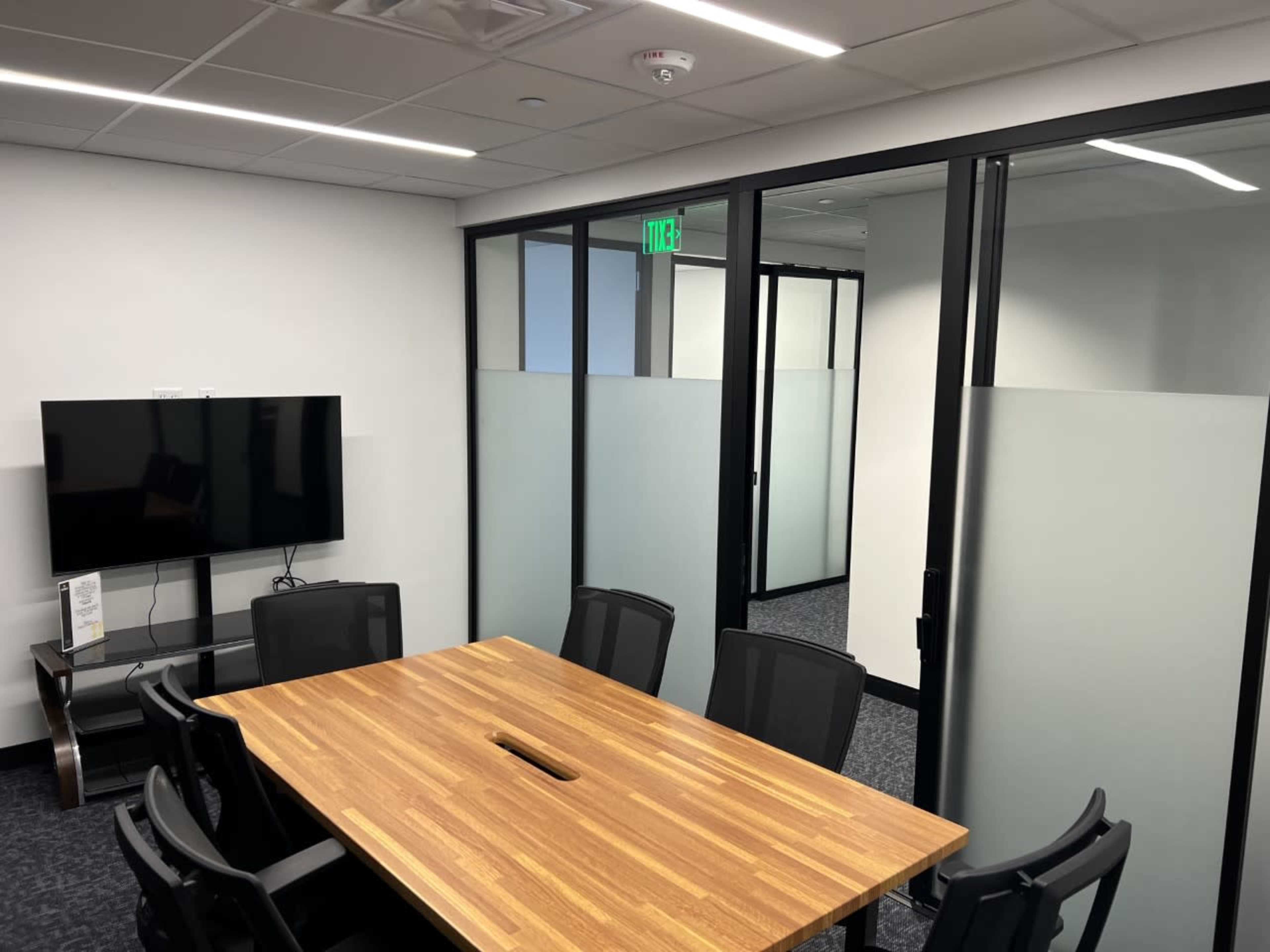A modern conference room with a wooden table, black office chairs, a television mounted on the wall, and glass partitions.