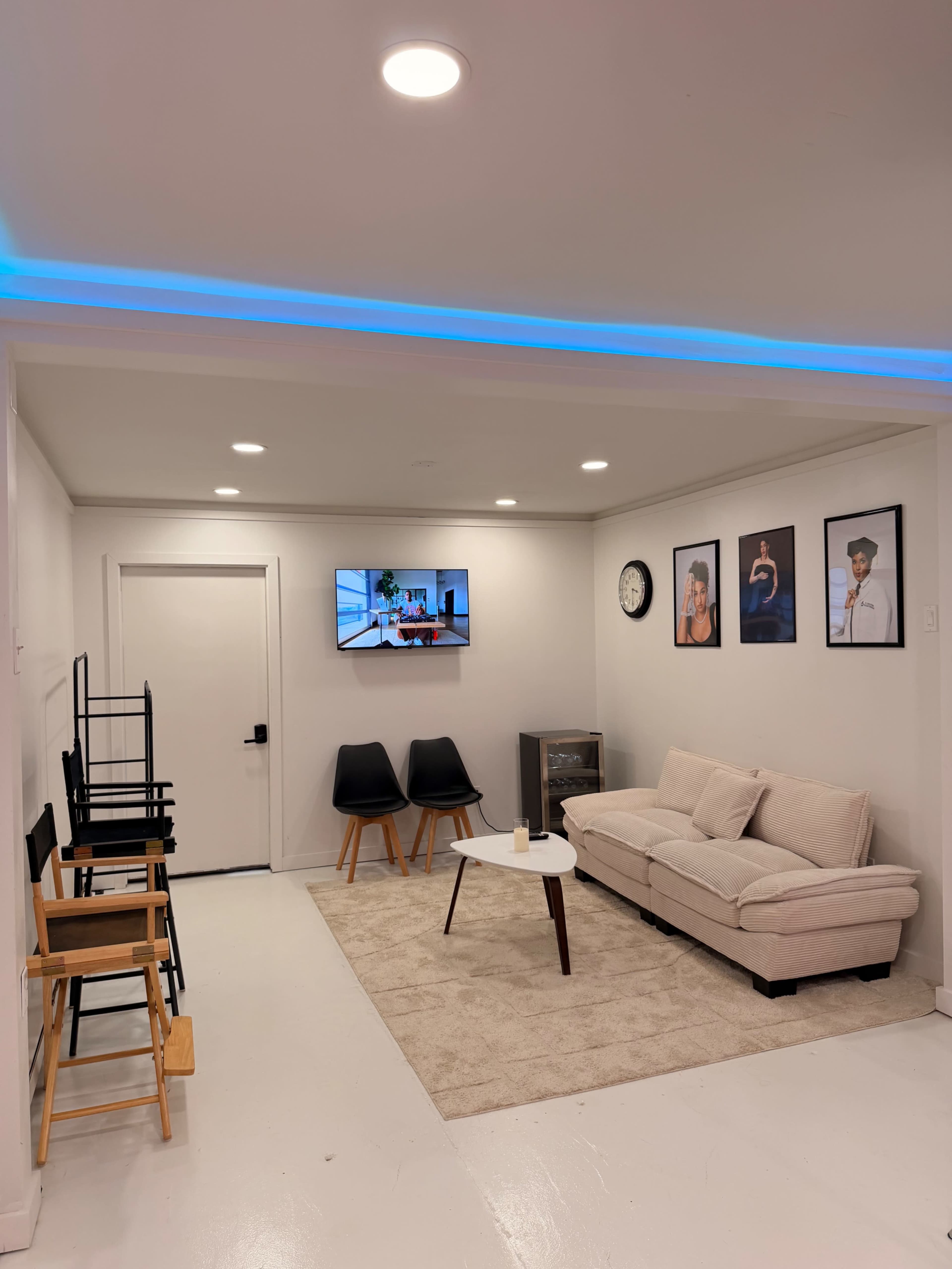 The image shows a modern indoor space featuring a beige sofa, two black chairs, a coffee table, and a wall-mounted television, with framed artworks and a clock adorning the walls.