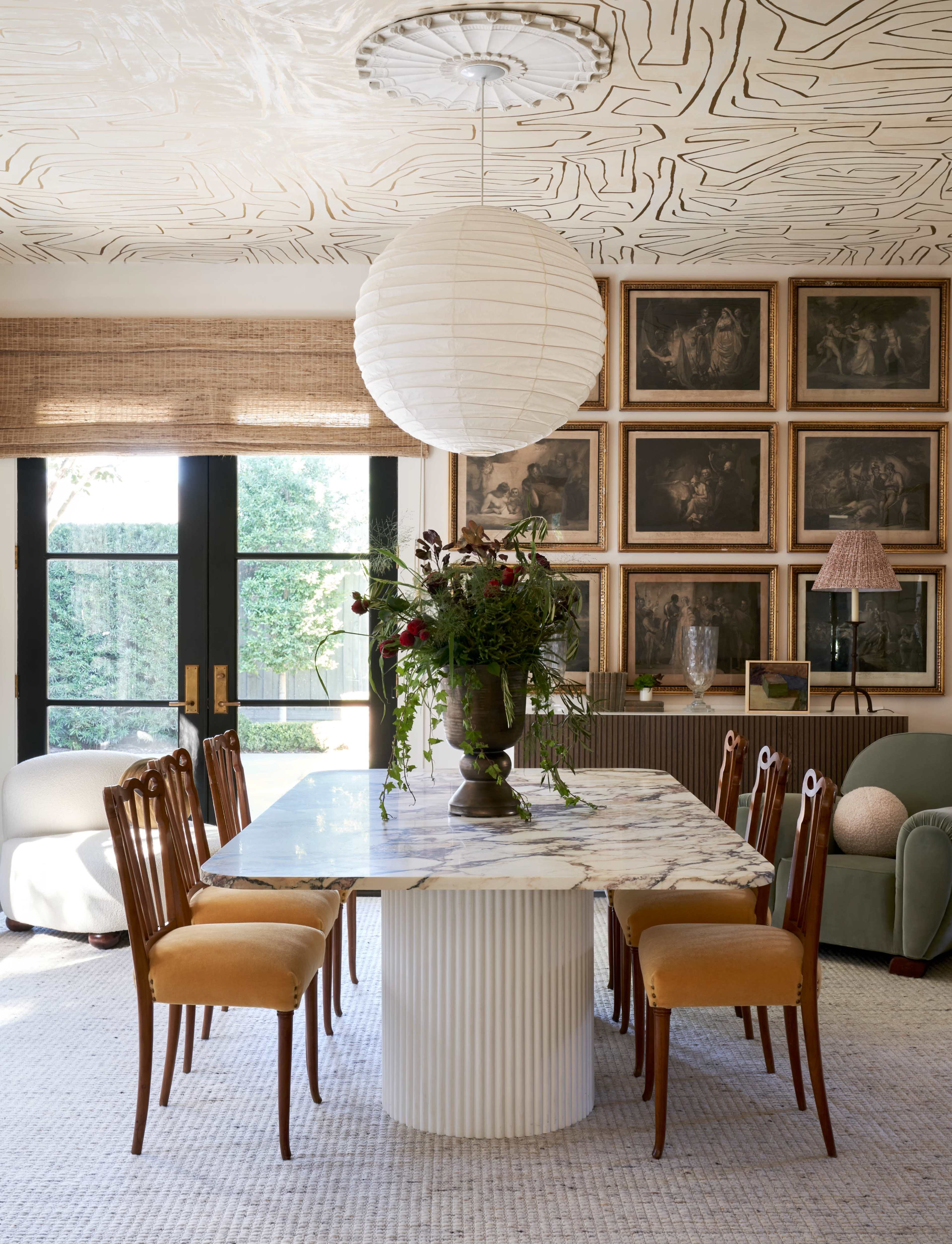 A dining area features a large marble table with eight wooden chairs, surrounded by framed artwork and illuminated by a pendant light.