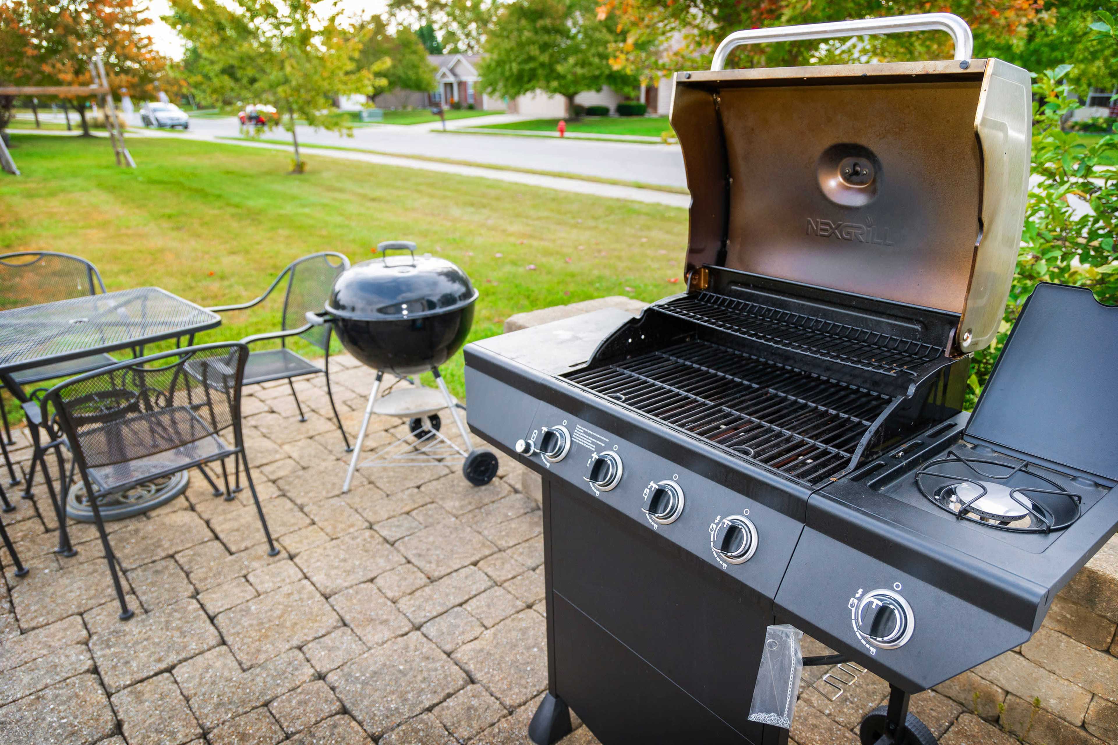 A gas grill with its lid open next to a charcoal grill, set on a stone patio with a table and chairs in a green yard.