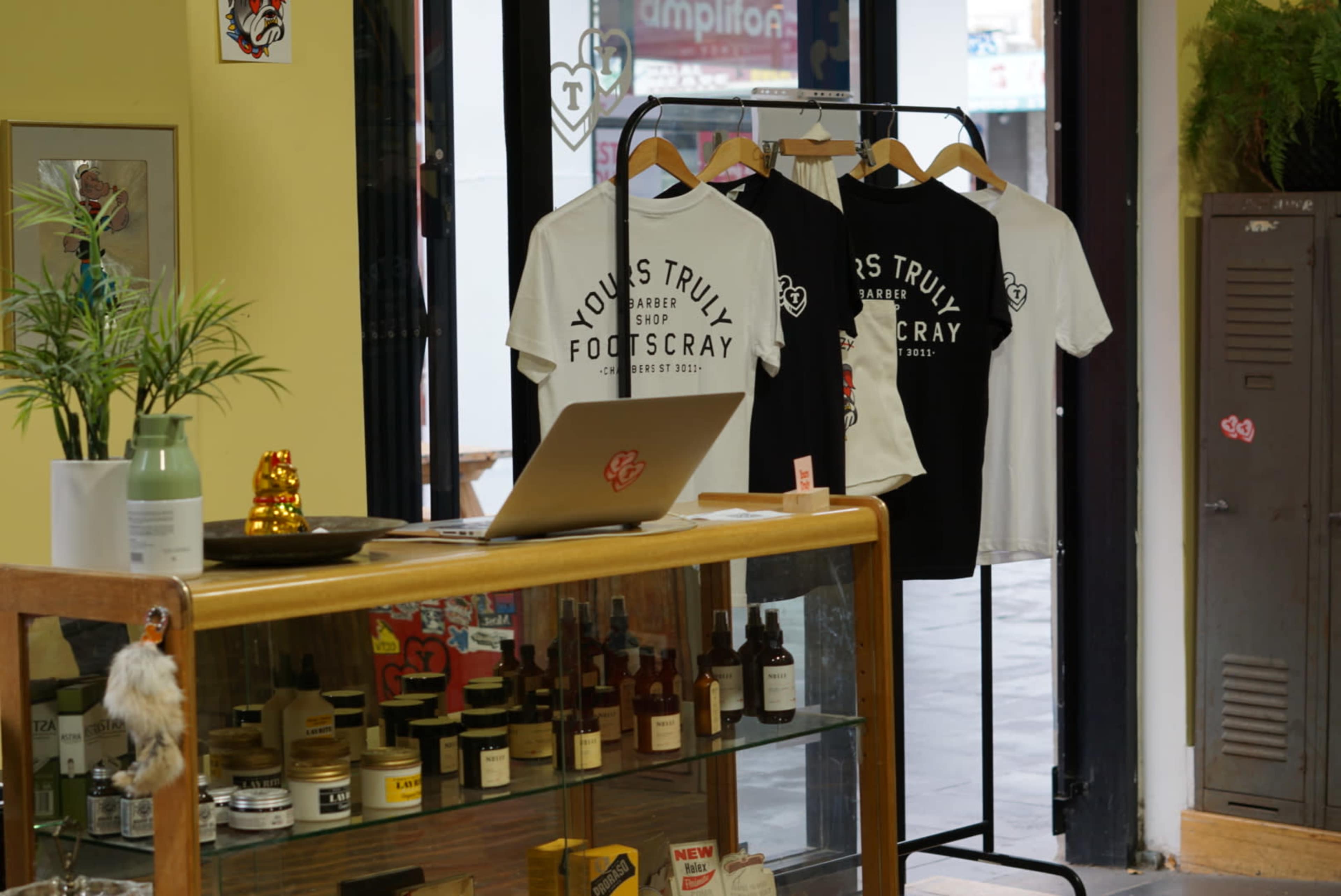 A retail space featuring a display of T-shirts on a rack, a wooden counter with various bottles, and a laptop on top.