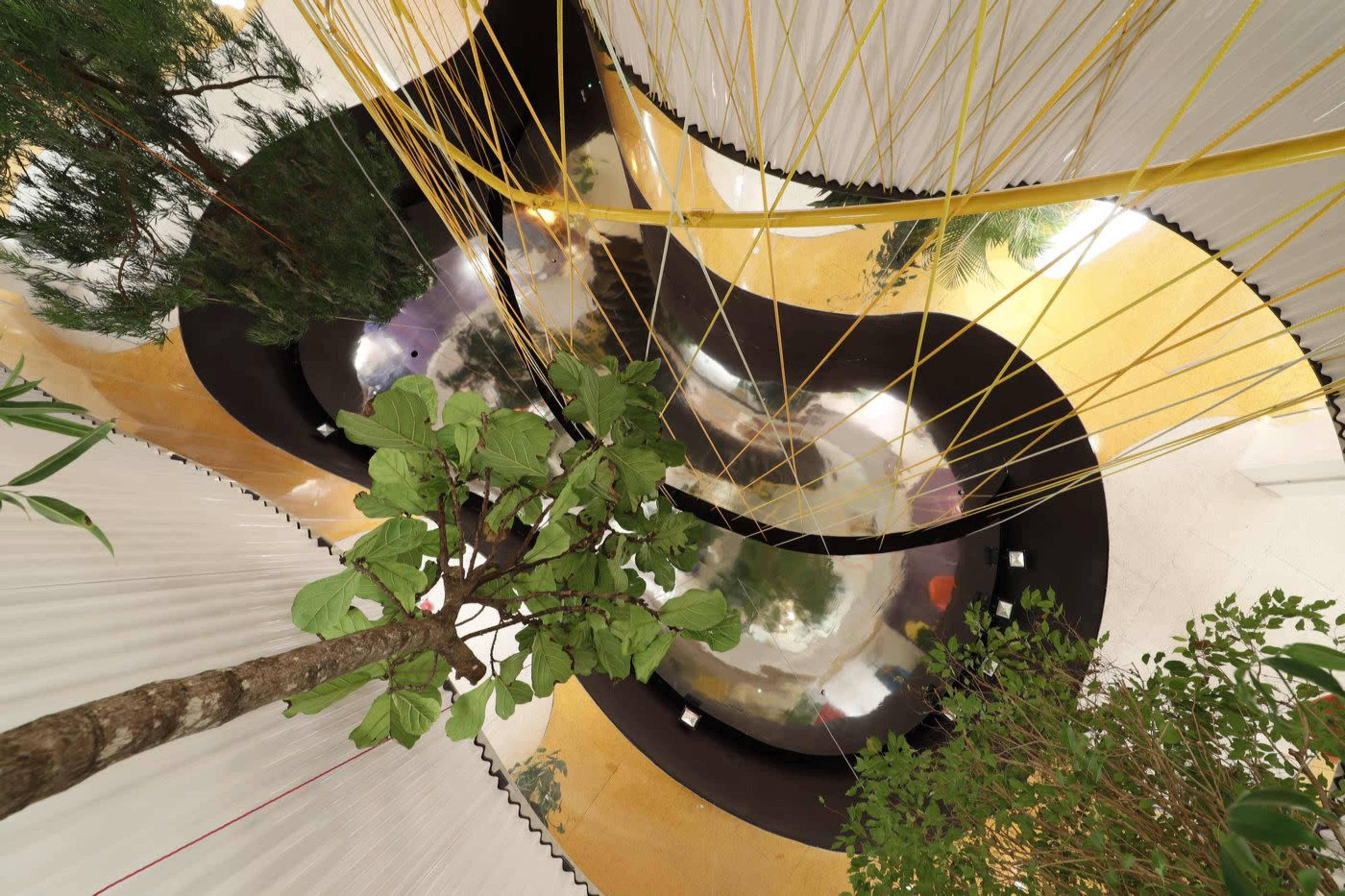 A modern, curved interior space featuring reflective surfaces, yellow lines hanging from the ceiling, and several indoor plants.