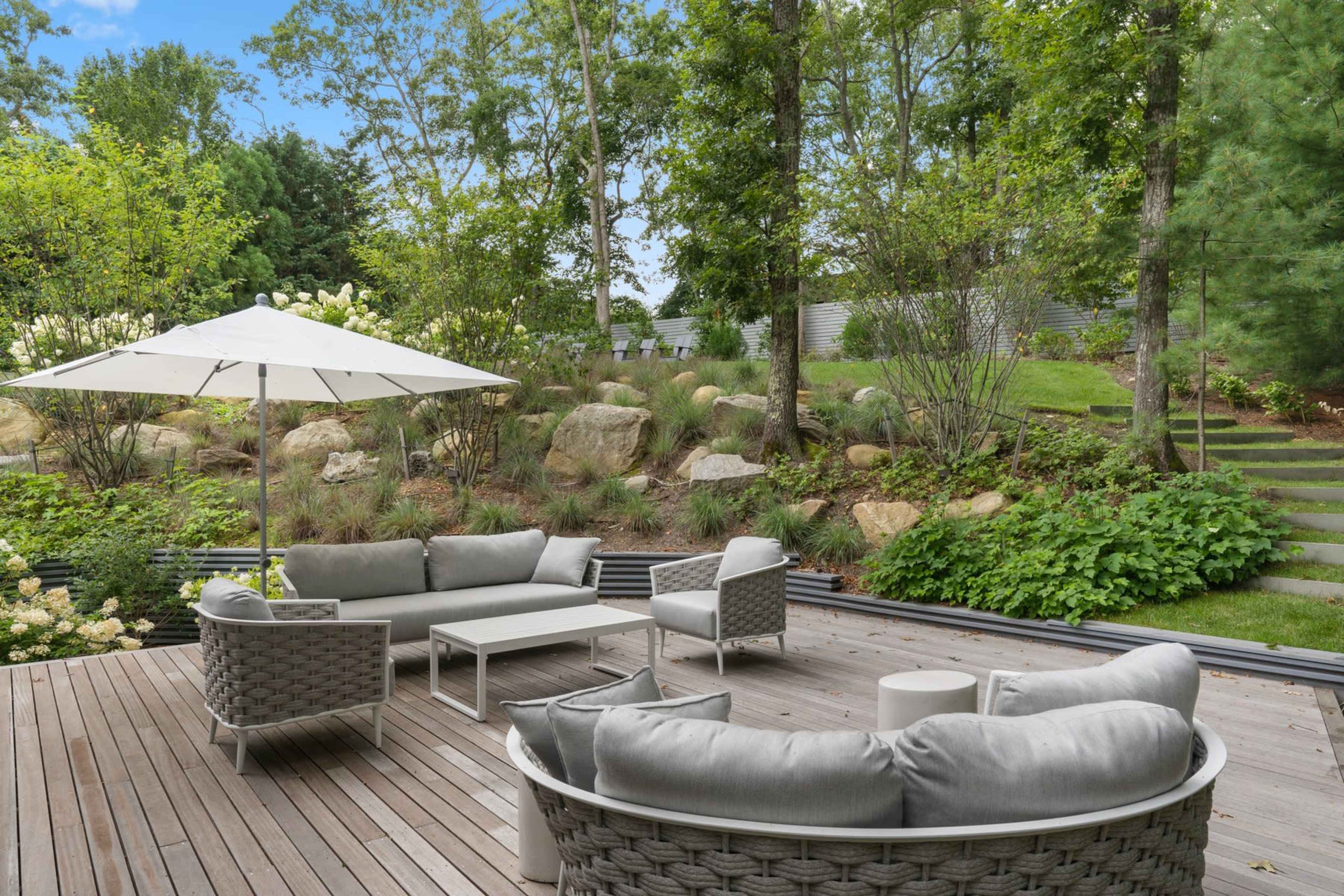 A patio area features circular gray wicker seating arrangements, a small table, and a white umbrella, surrounded by greenery and stone landscaping.