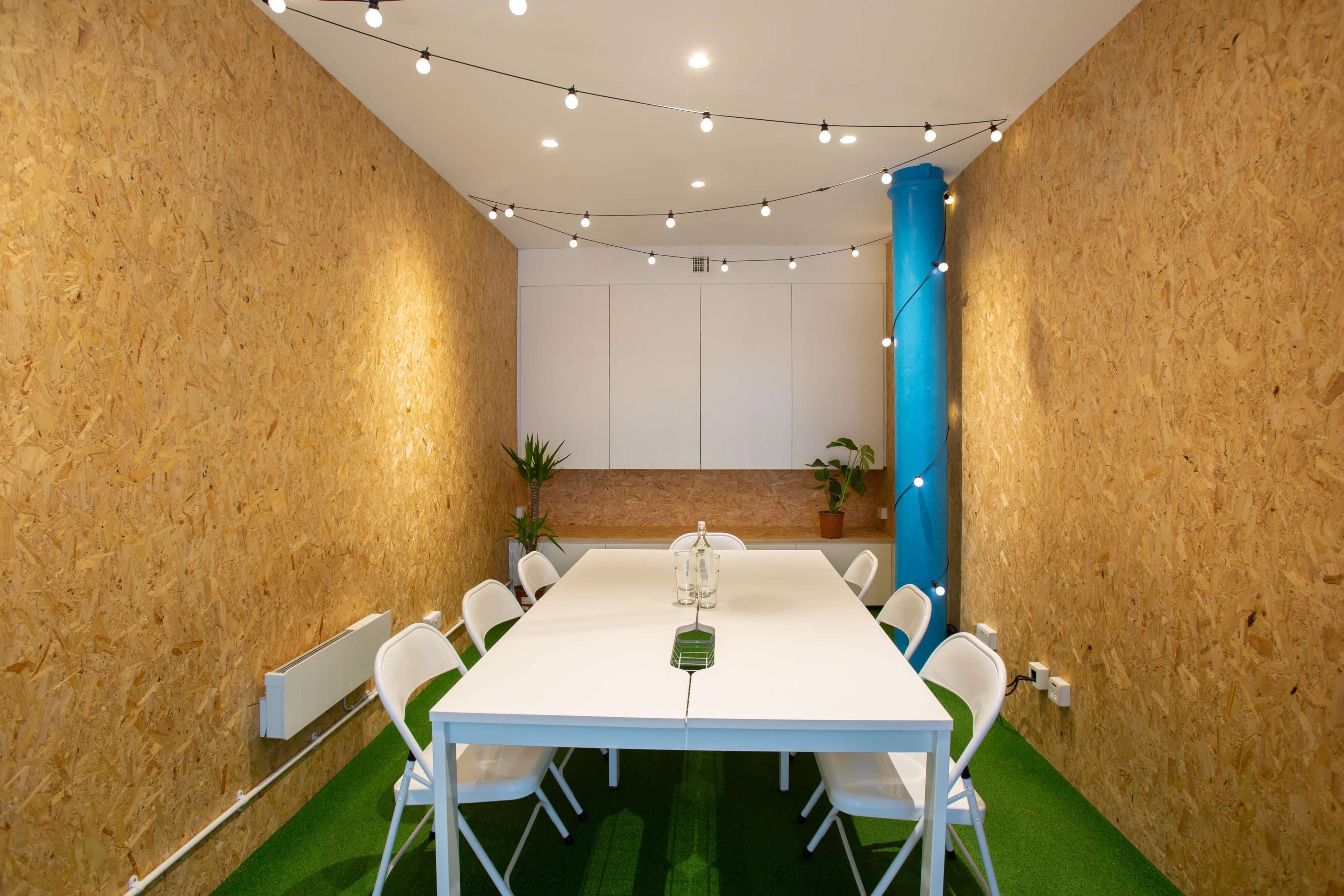 A meeting room features a rectangular white table surrounded by eight white chairs, with wooden walls and a green carpet, illuminated by string lights overhead.