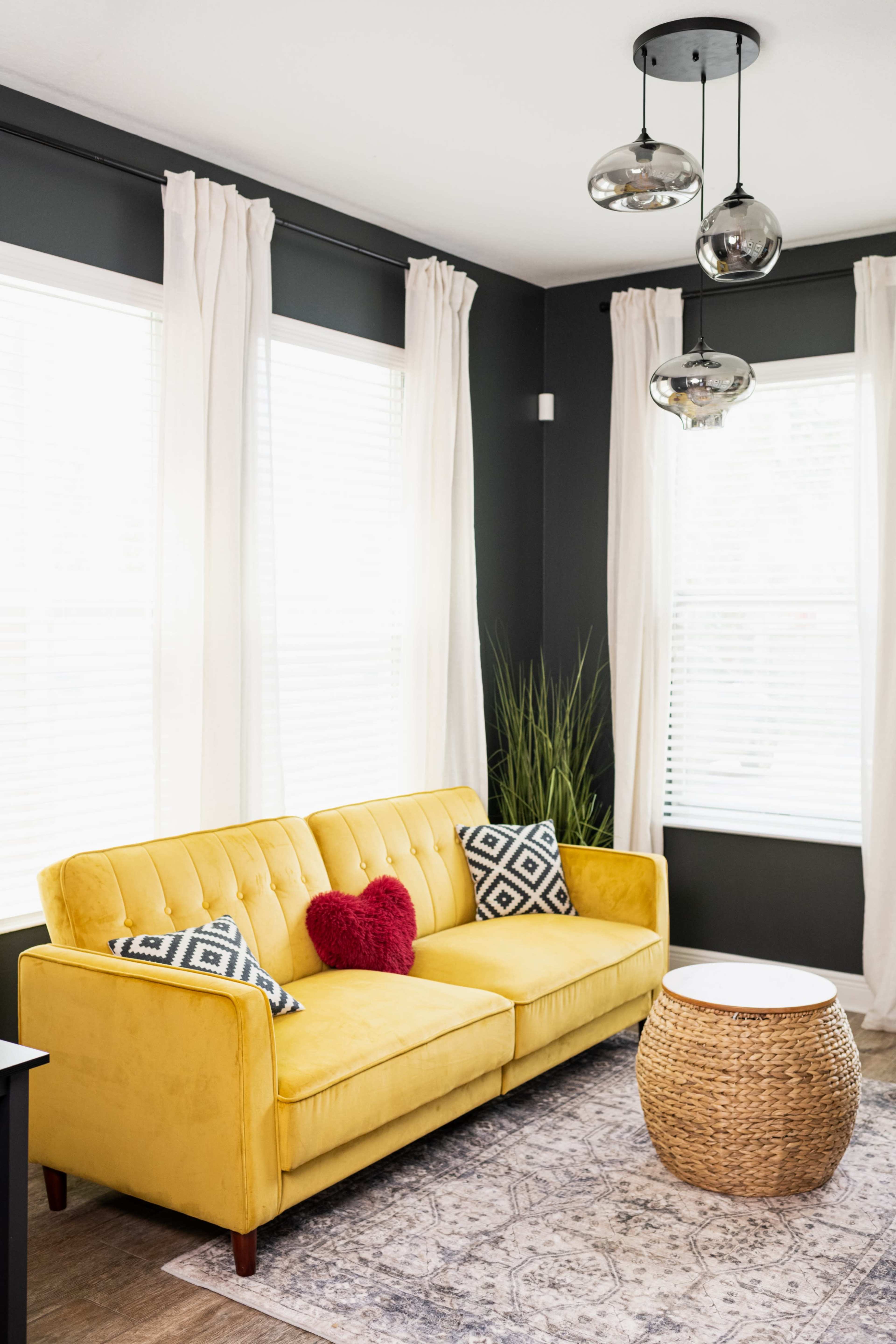 A yellow couch with patterned throw pillows sits against a dark wall, surrounded by large windows with white curtains and a round wicker table.