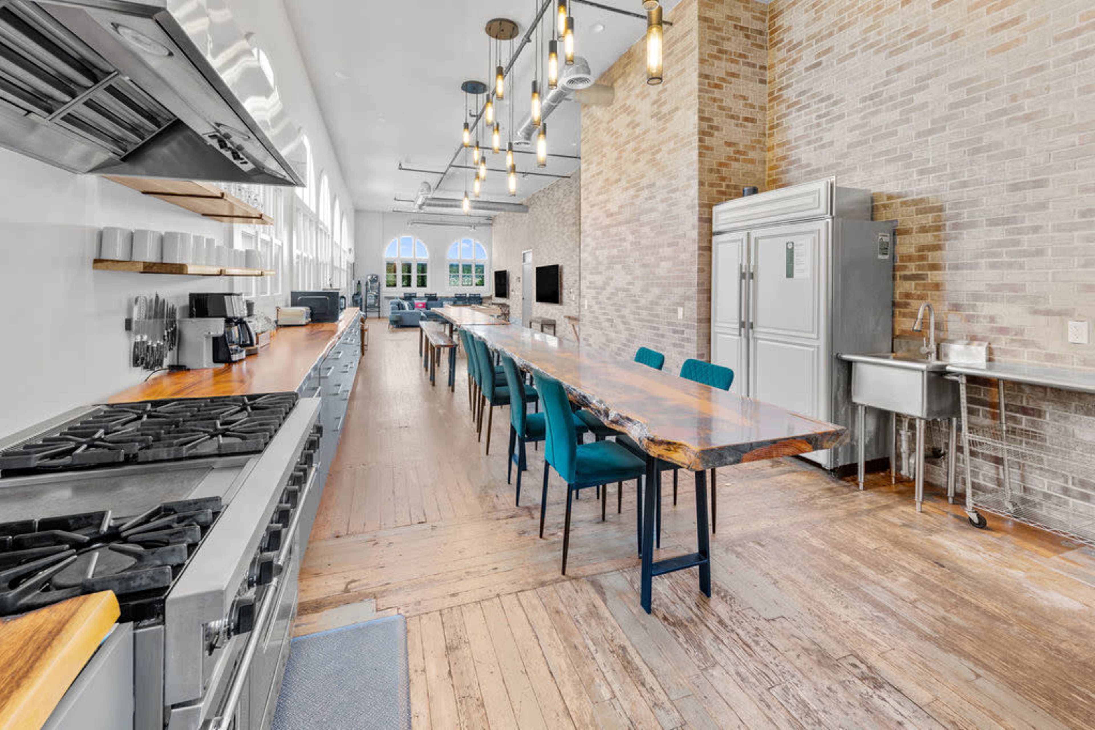 The image shows a spacious kitchen with a long wooden table, industrial-style appliances, and large windows that allow natural light to illuminate the space.