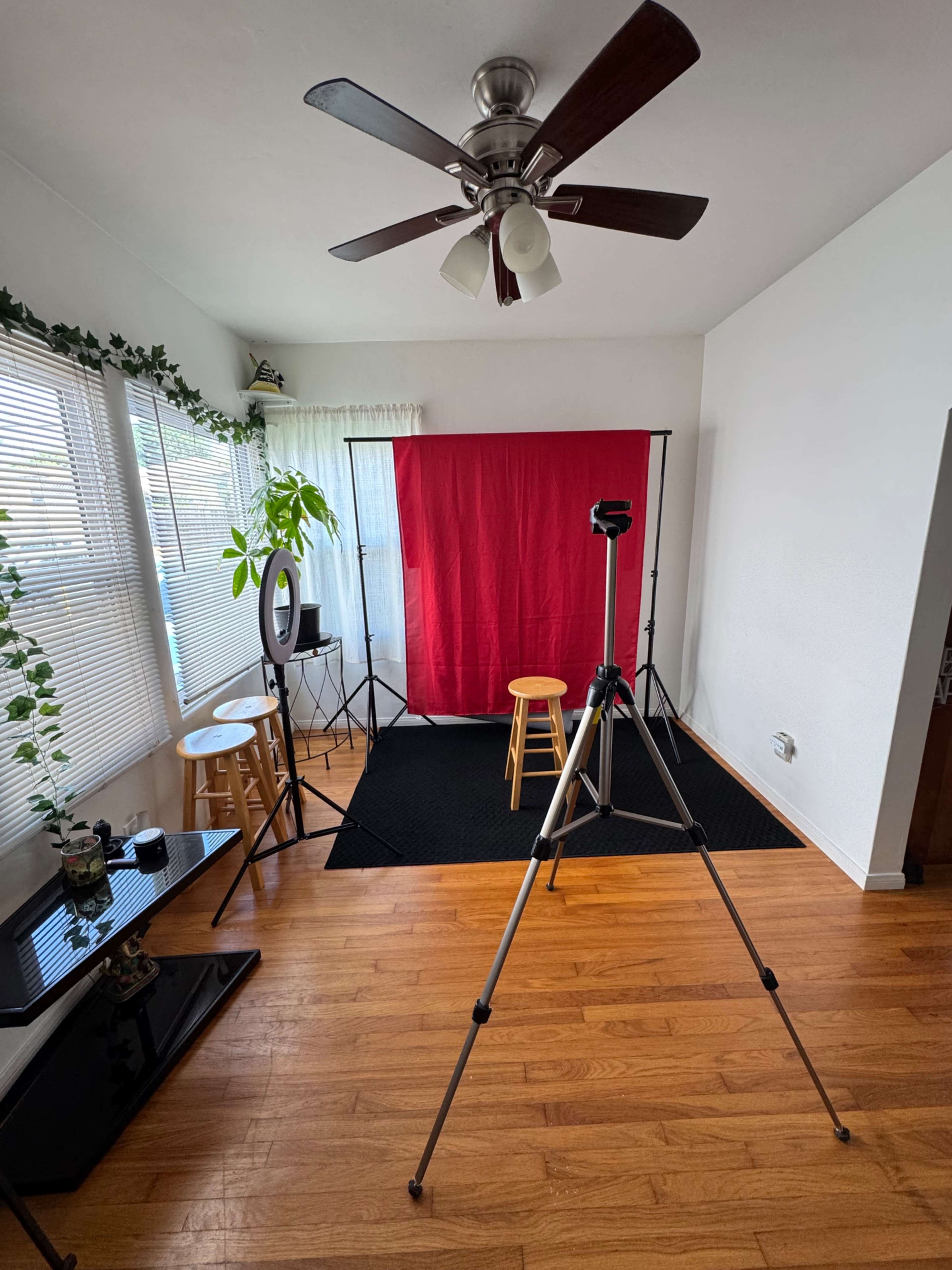 The image shows a small studio setup with a red backdrop, a camera on a tripod, and seating arranged for a video or photo shoot.