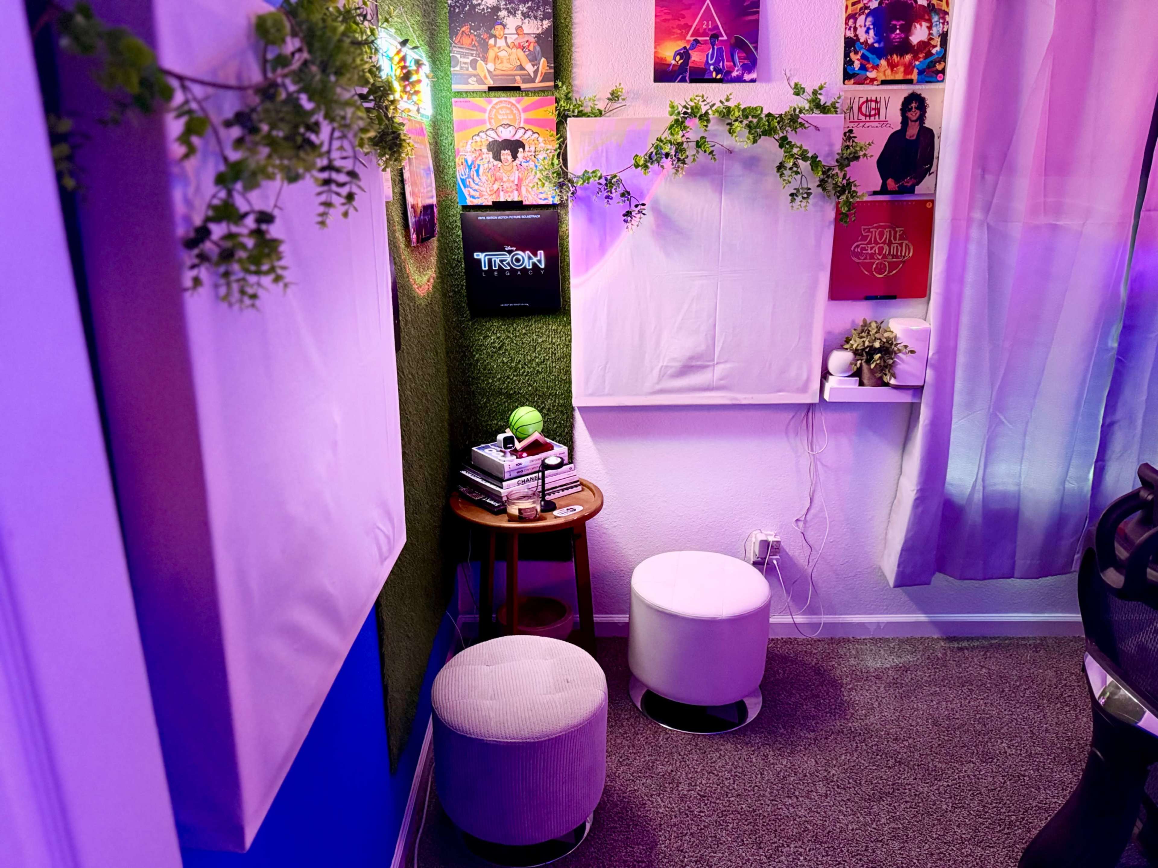 The image shows a cozy corner of a room featuring two white stools, a small table with books, and posters on the wall, all illuminated by colorful lighting.