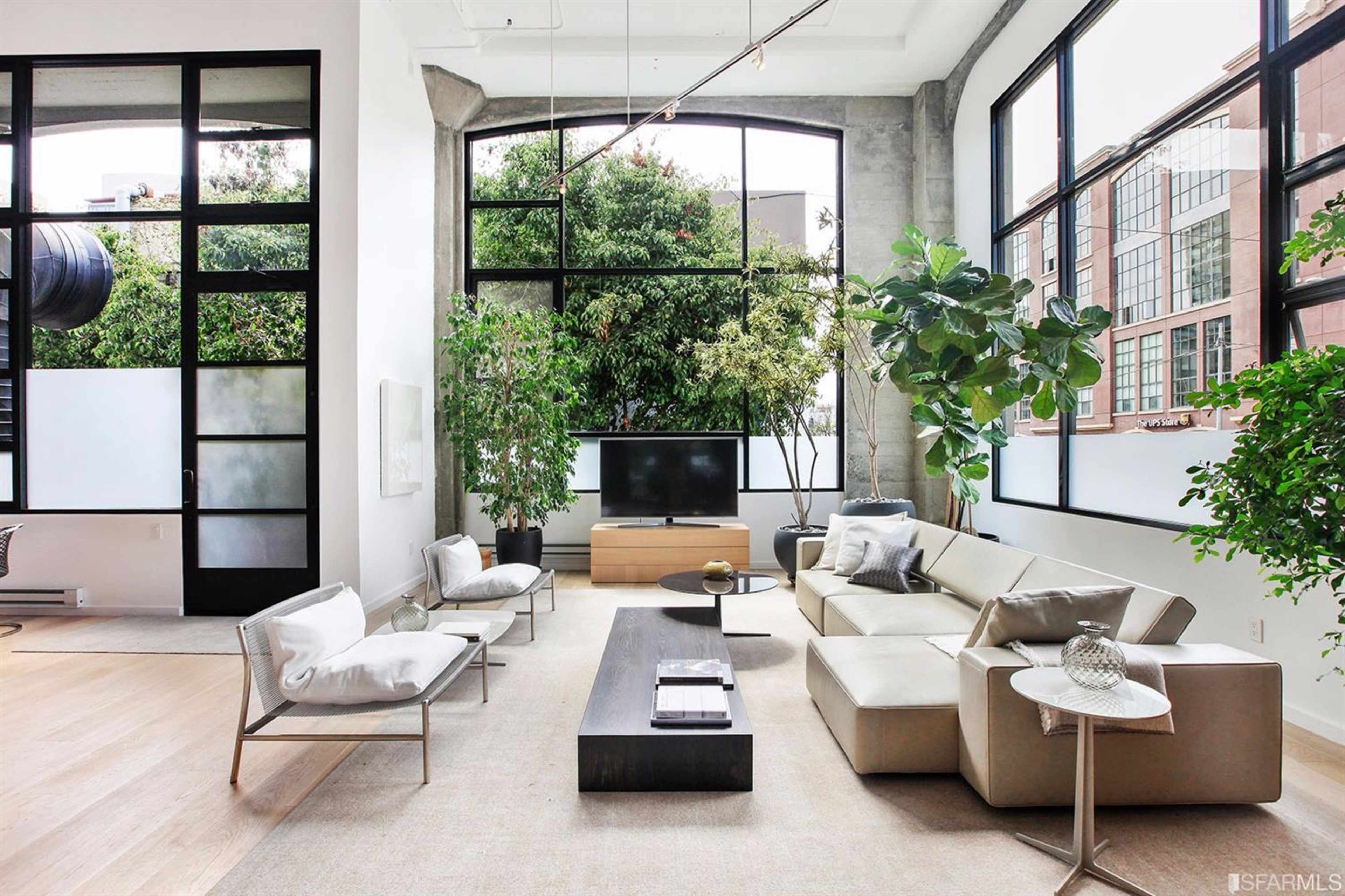 A modern living room with large windows, a television, a beige sectional sofa, plants, and minimalist furniture.