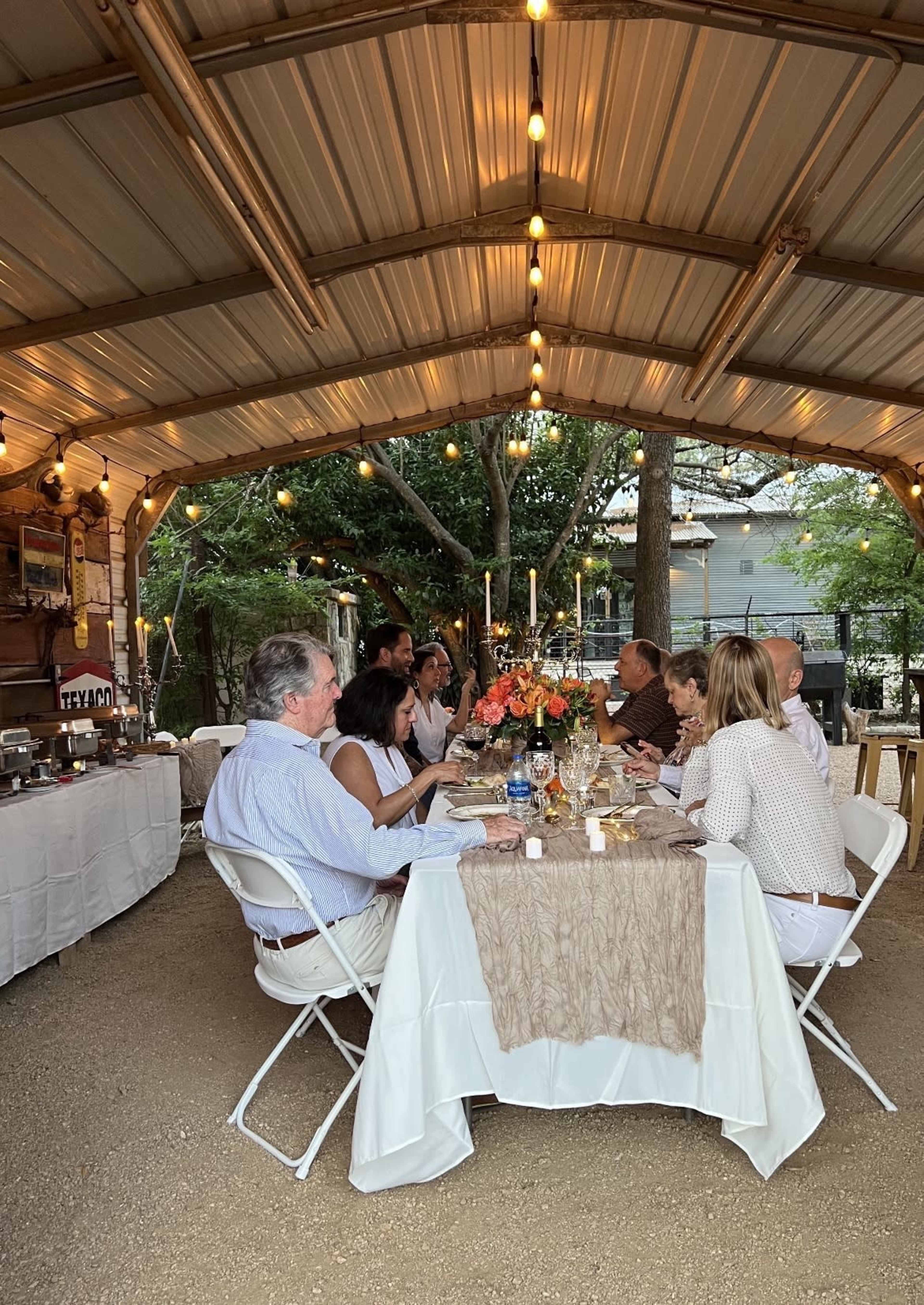 A group of people sits around a table decorated with flowers and candles in a covered outdoor dining area.