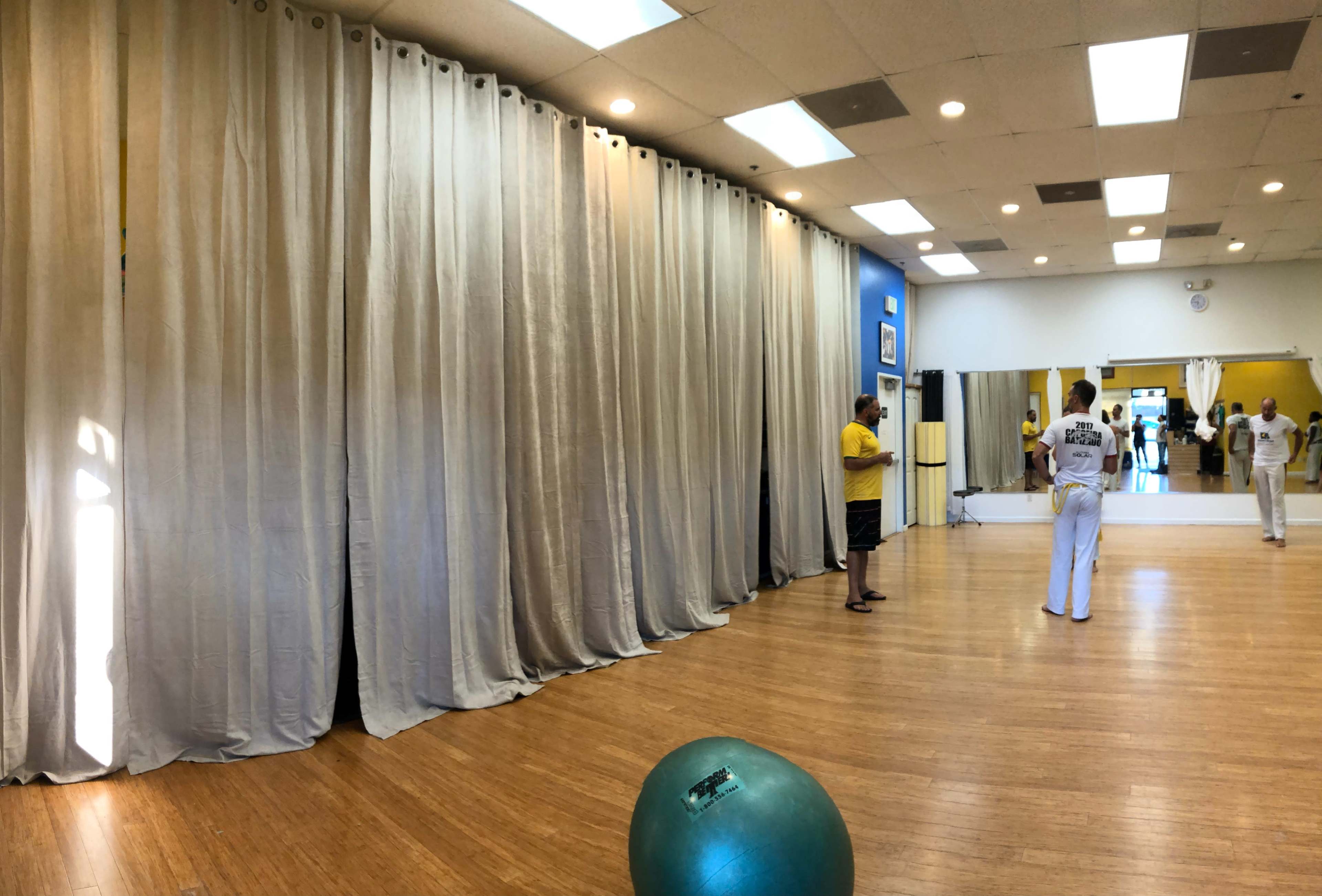 A spacious studio with wooden flooring, large curtains, a green exercise ball, and a few people engaging in activities in the background.