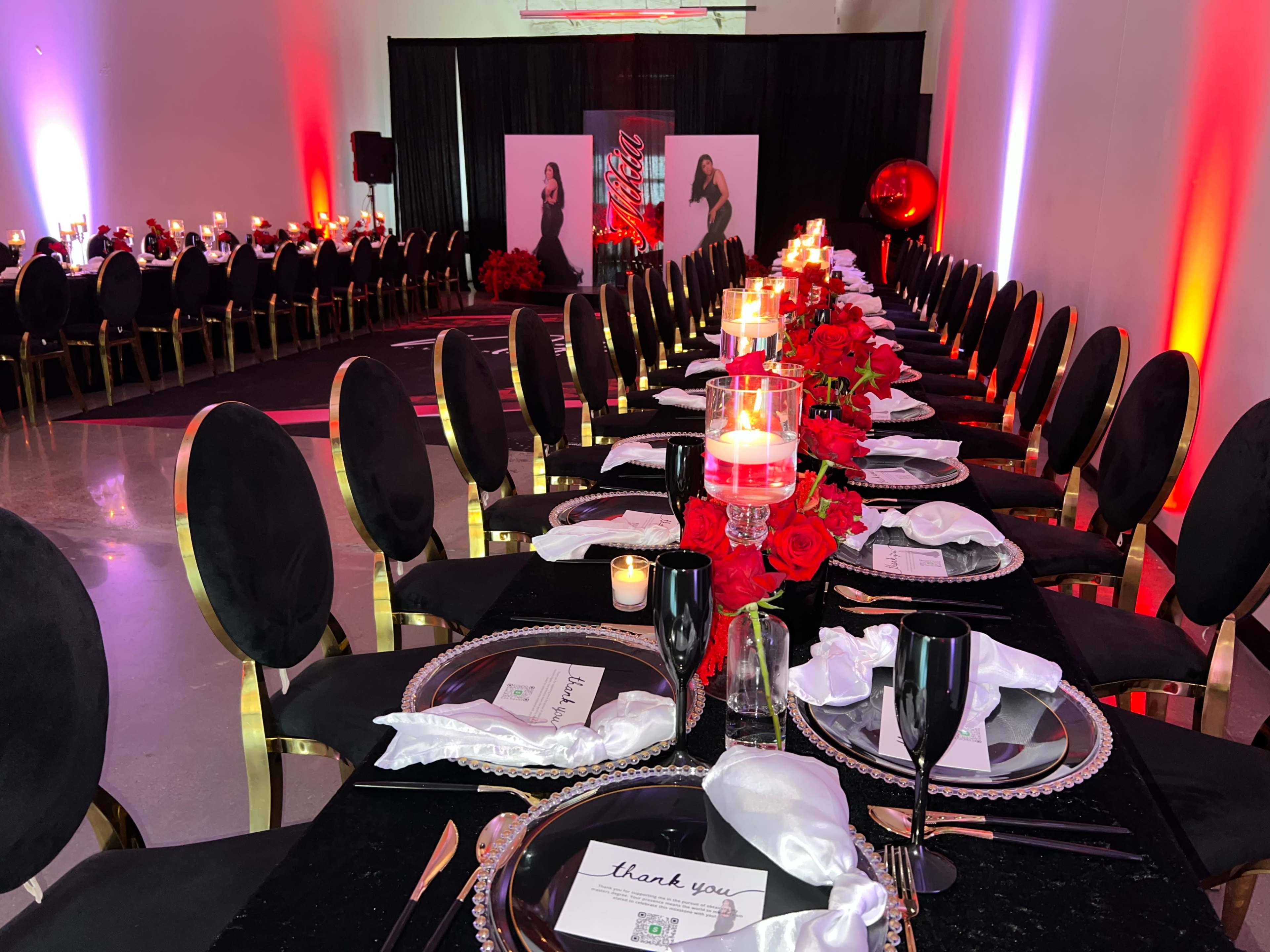 A long banquet table is elegantly set with black placemats, white napkins, and candles, surrounded by gold-accented chairs in a dimly lit event space.