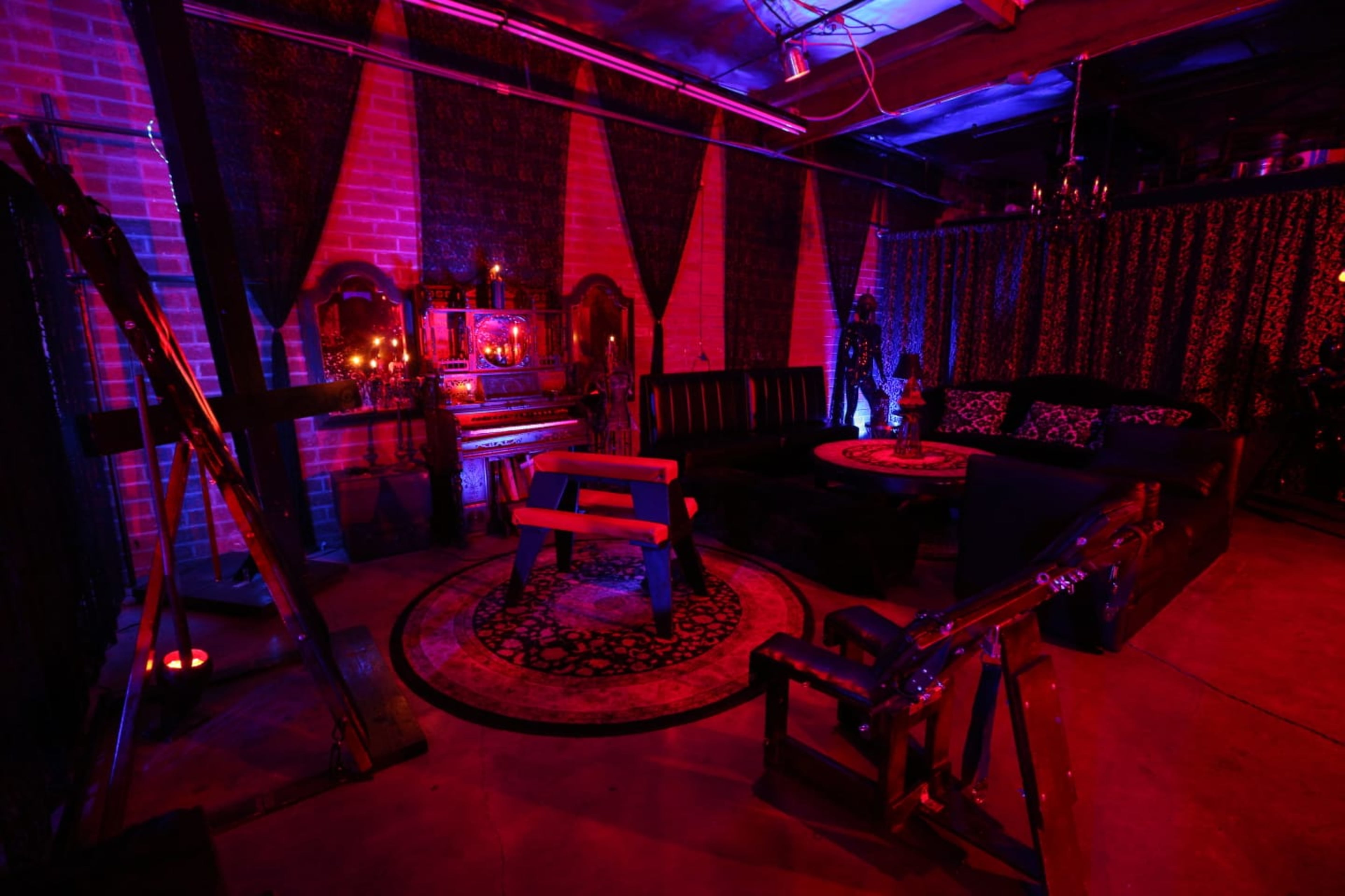 A dimly lit room with a black sofa, a wooden table, a vintage piano, and various decorative elements against a backdrop of patterned fabric.