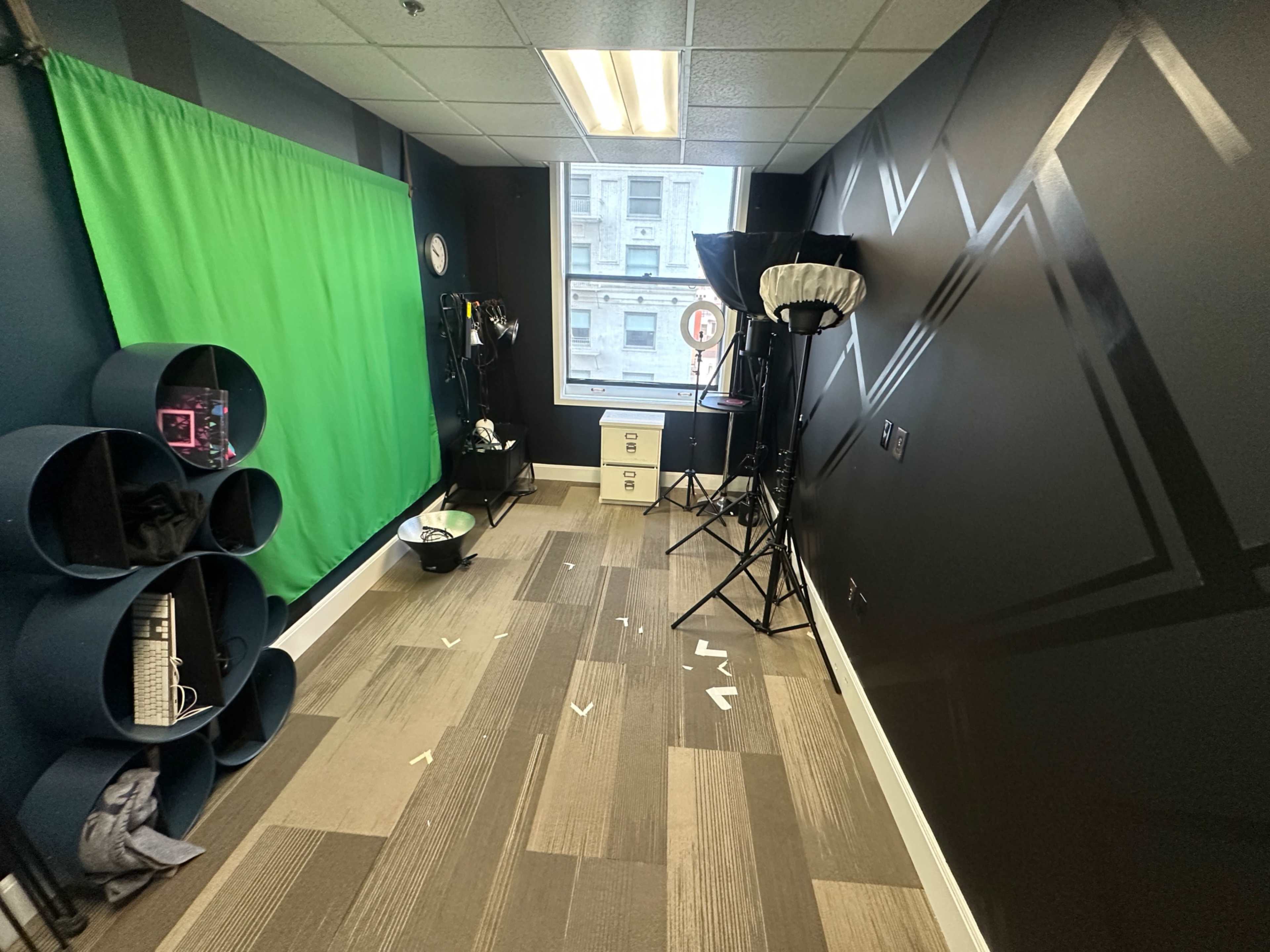 The image shows a small studio space with a green screen, lighting equipment, and a desk with storage drawers.