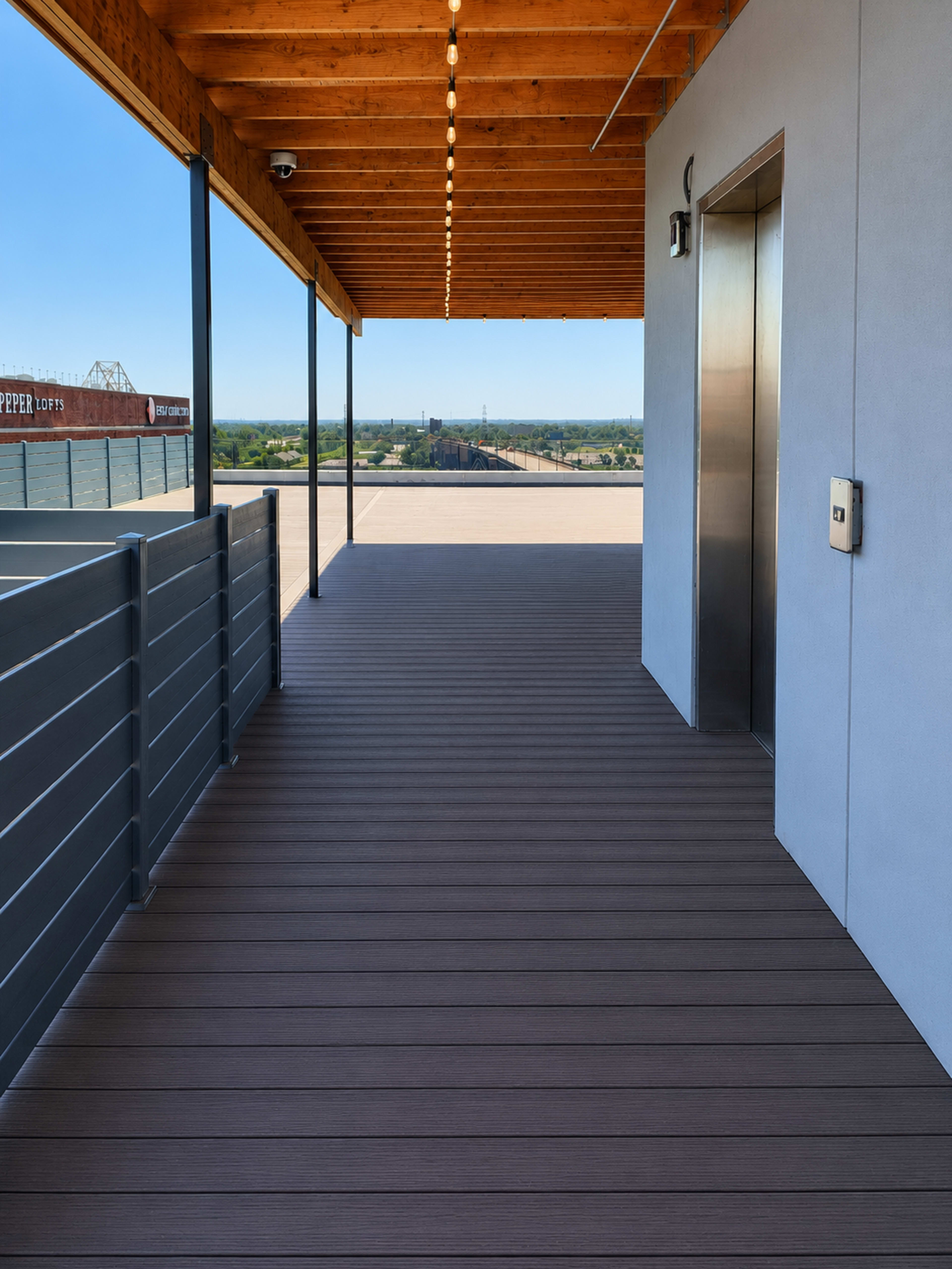 The image shows a modern outdoor corridor with wooden ceiling beams, an elevator door, and a railing overlooking a vast landscape.