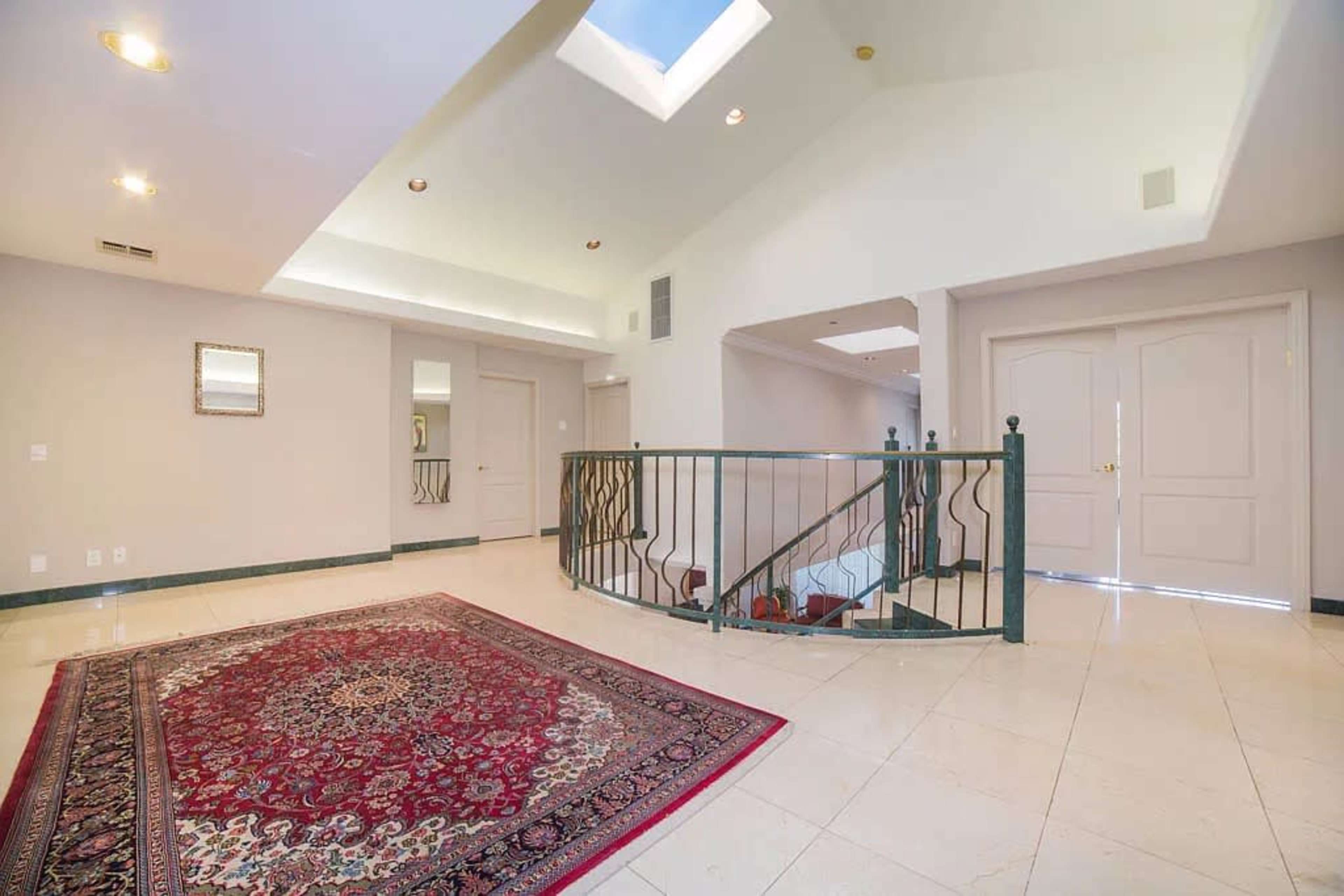 The image shows a spacious foyer with a decorative rug, a staircase with a wrought iron railing, and a skylight allowing natural light to enter the area.