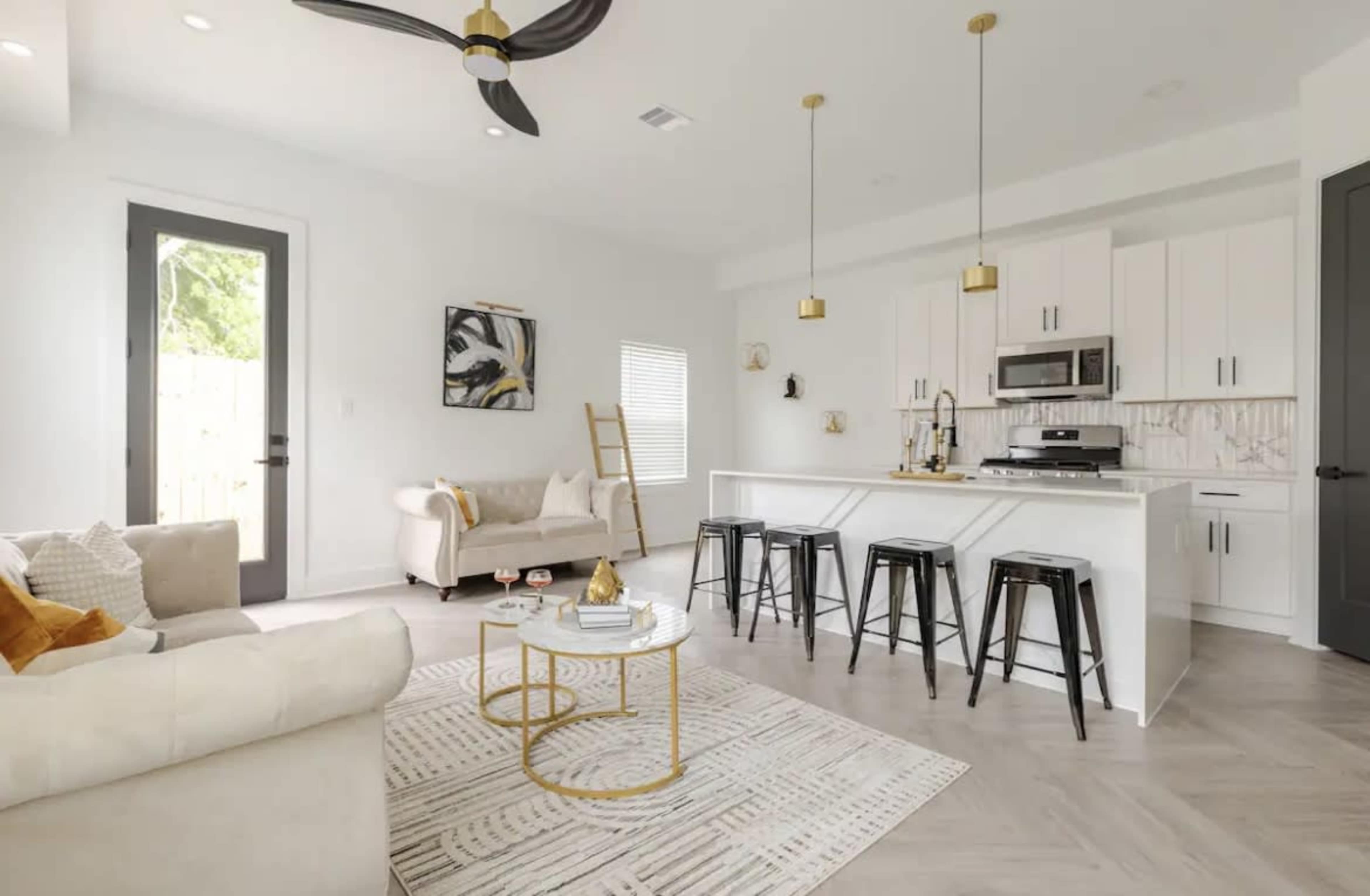A bright living space features a kitchen island with bar stools, a sofa, and a ladder against the wall, with large windows allowing natural light in.