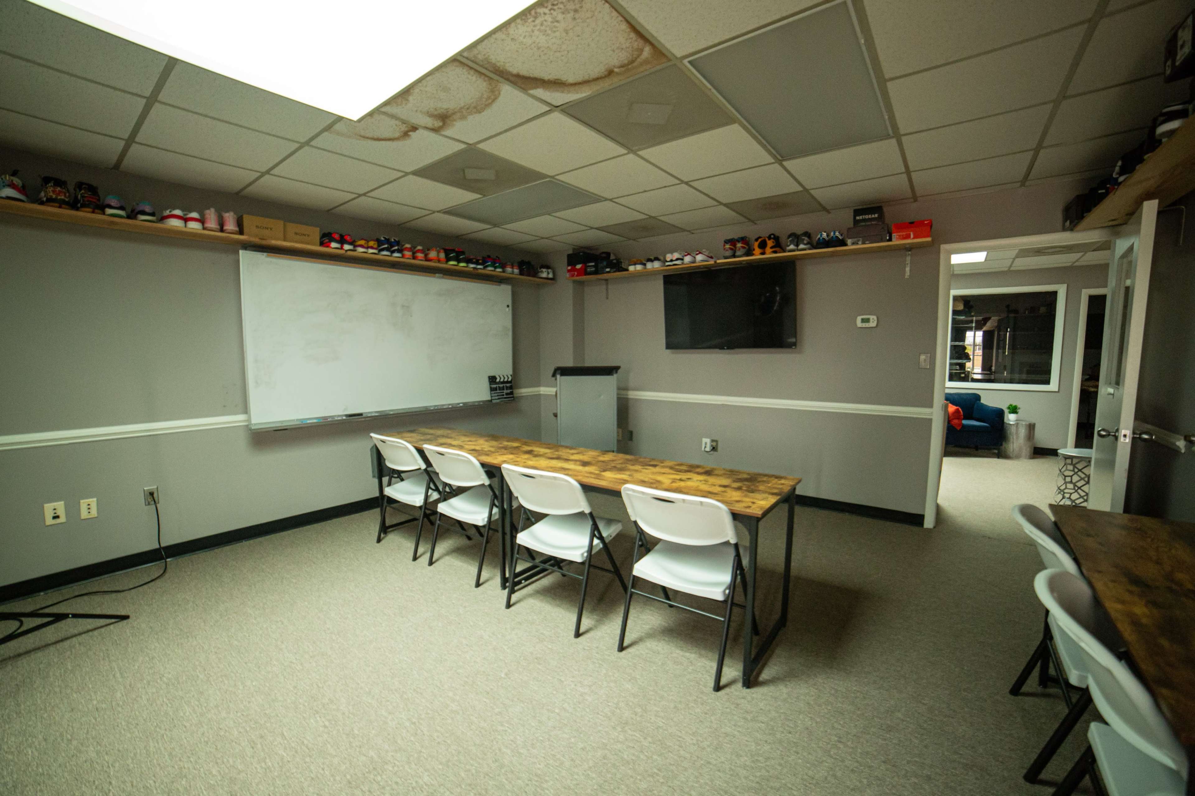 A meeting room with multiple tables and chairs, a whiteboard, a television, and shelves displaying various items.