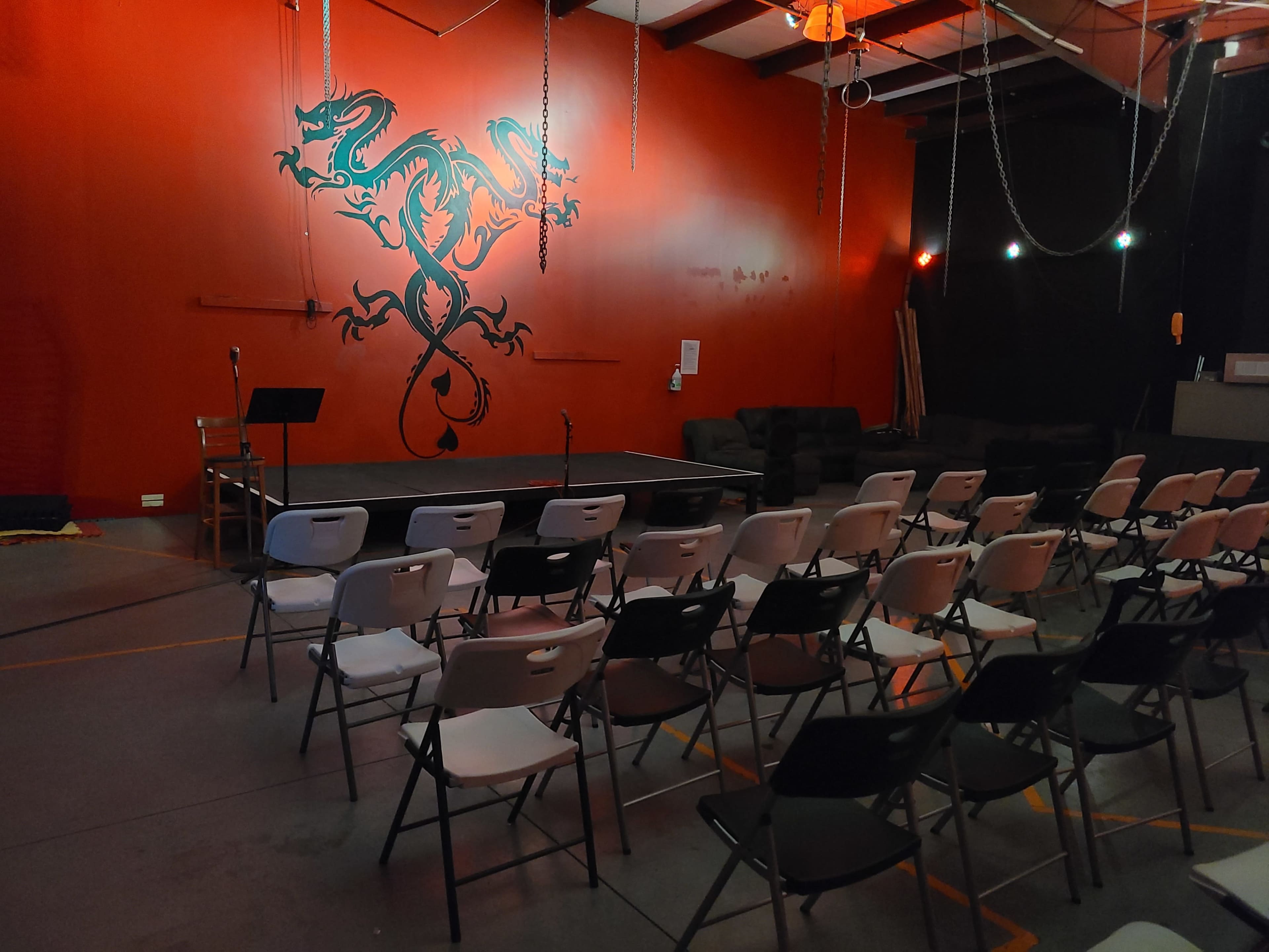 A performance space with a stage, rows of white and black chairs, and a large decorative dragon mural on an orange wall.