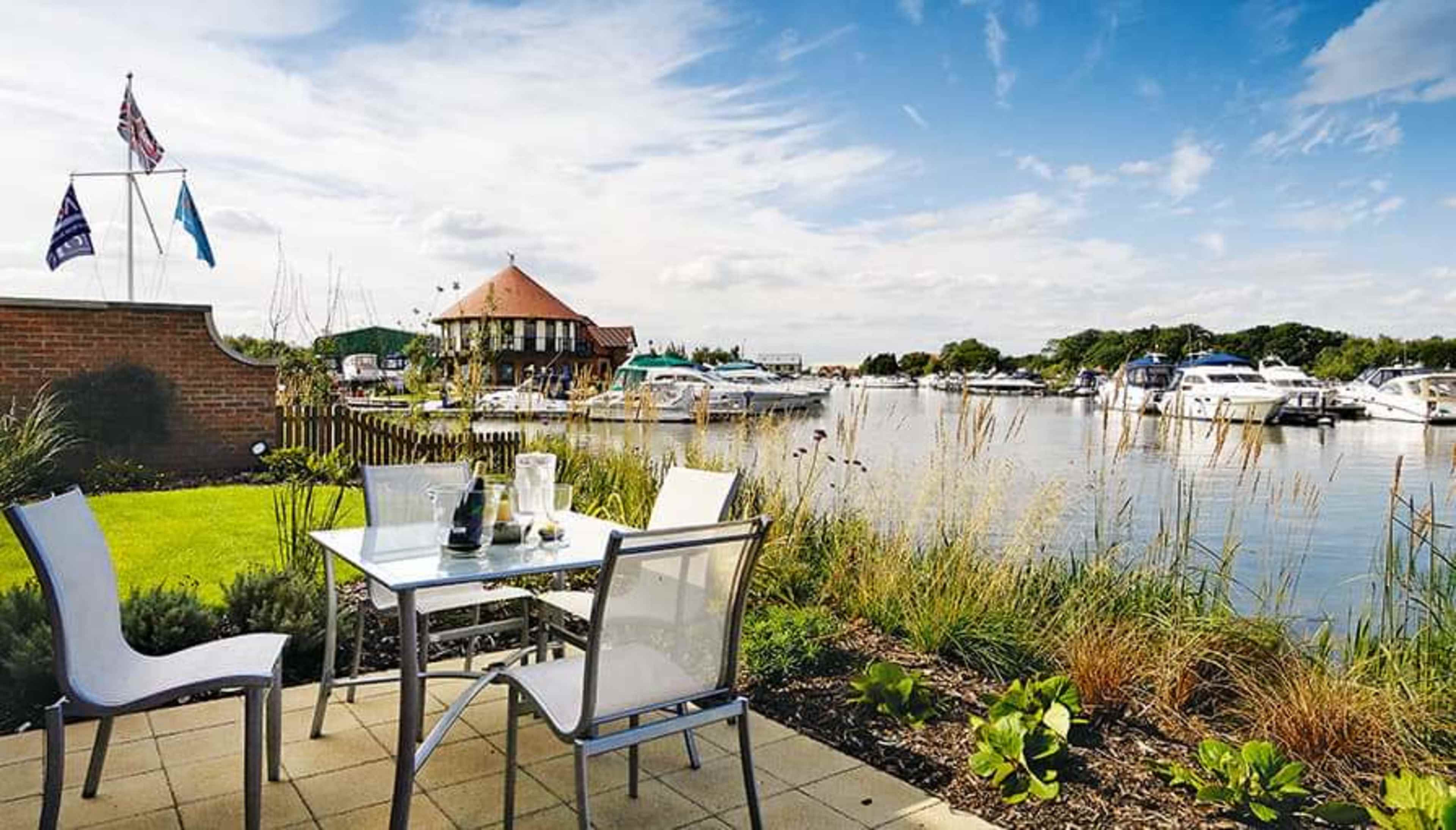 A patio with white chairs and a table overlooks a calm waterway filled with boats, framed by lush greenery and a building in the background.