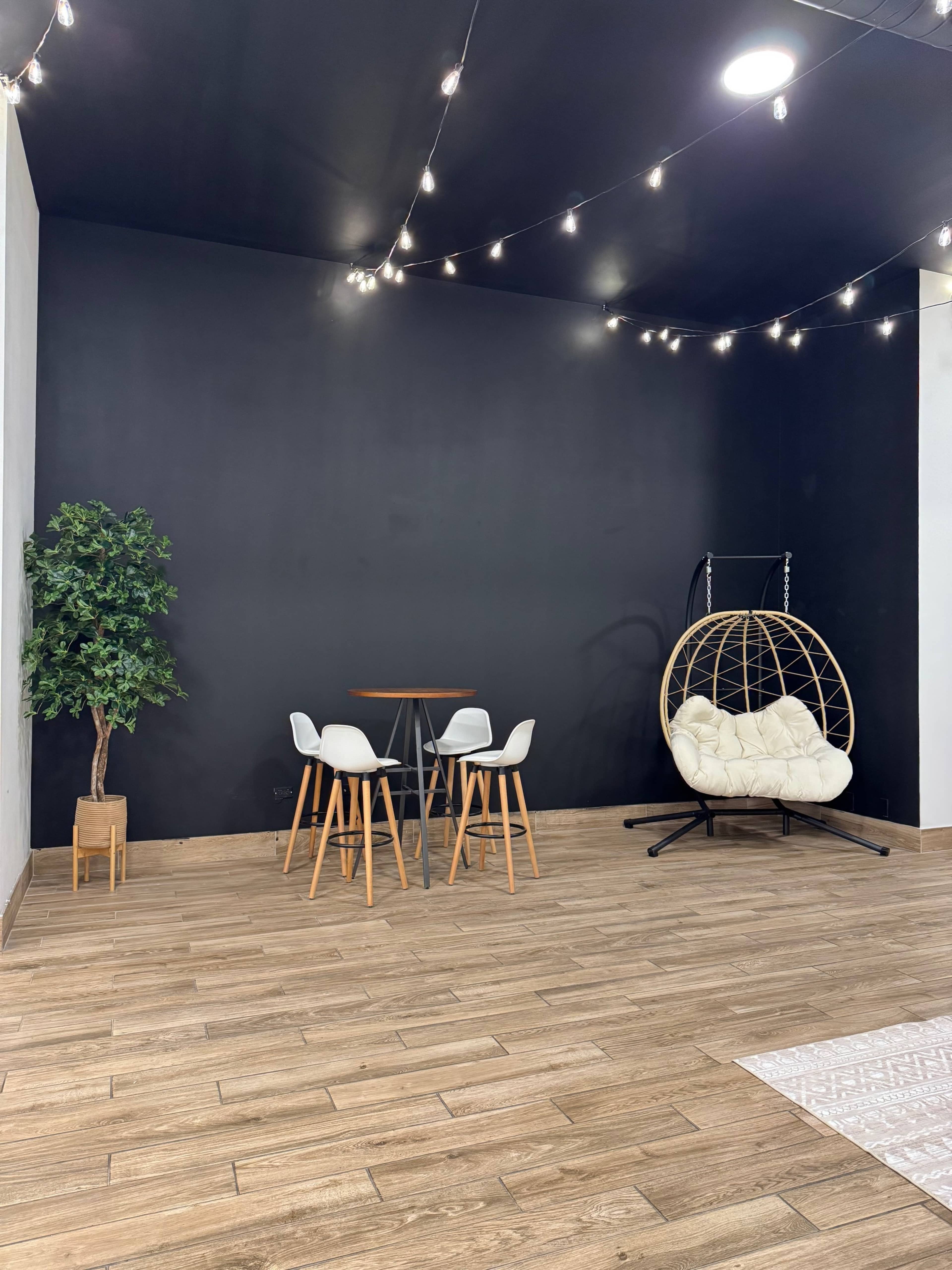 The image shows a minimalistic room with a black wall, a small round table and four chairs, a hanging chair, and a decorative plant.