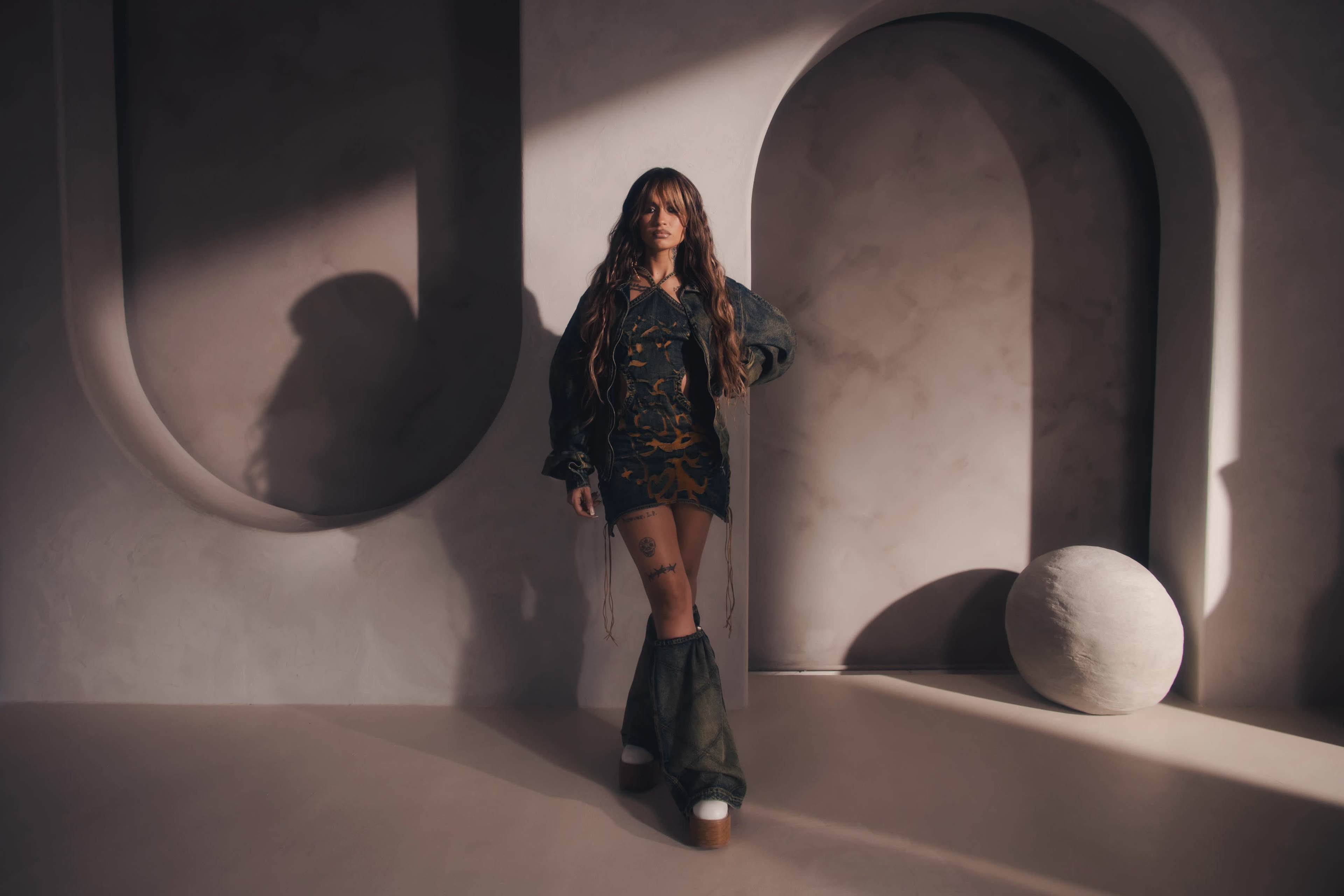 A person stands confidently in a stylish outfit against a textured backdrop with curved elements and a spherical object on the floor.