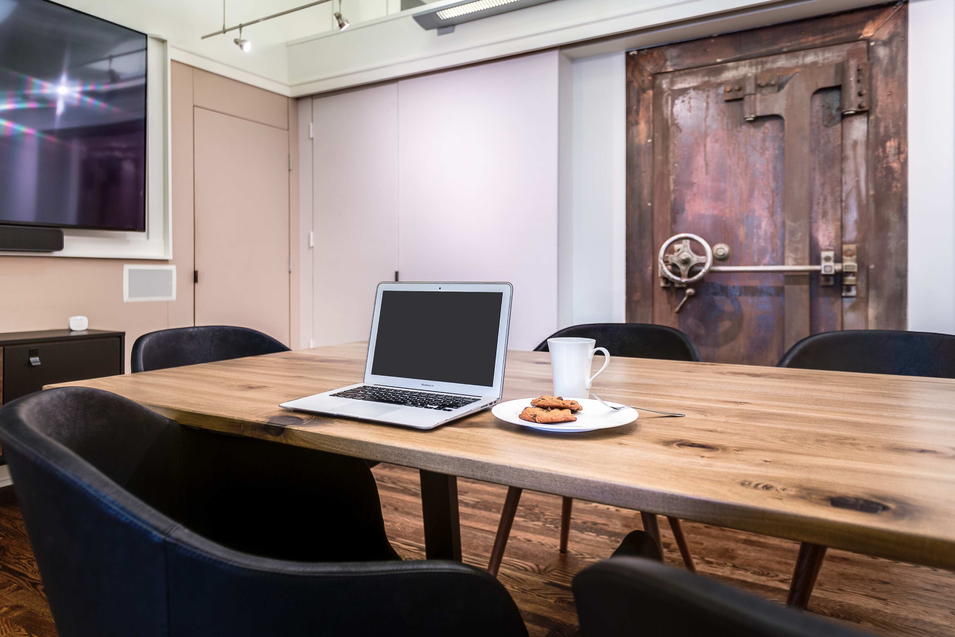 A laptop, a white cup, and a plate with a cookie are placed on a wooden conference table in a room with a brown door and dark chairs.