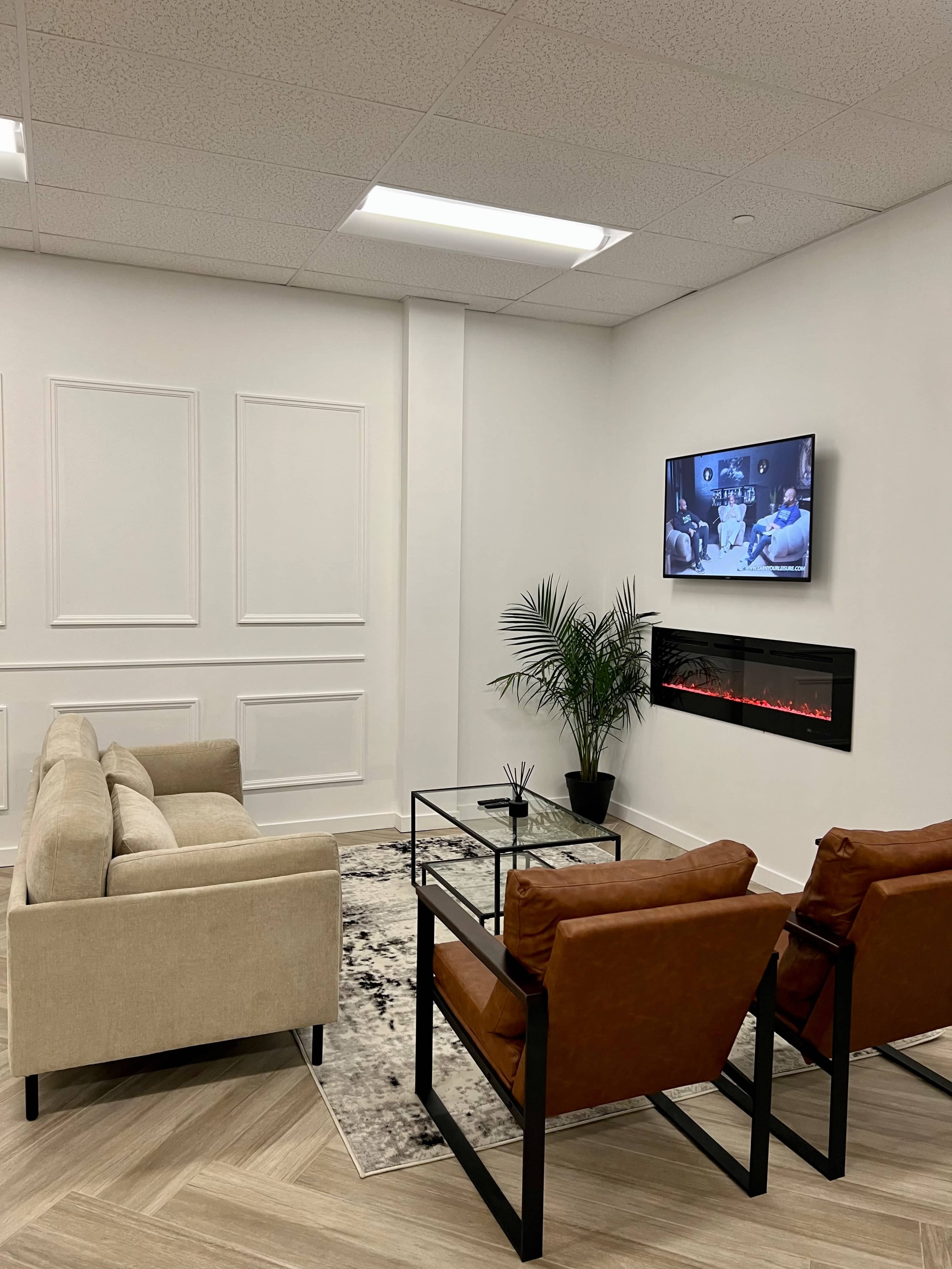 A modern living room with two brown armchairs, a beige couch, a glass coffee table, an electric fireplace, and a television mounted on a white wall.