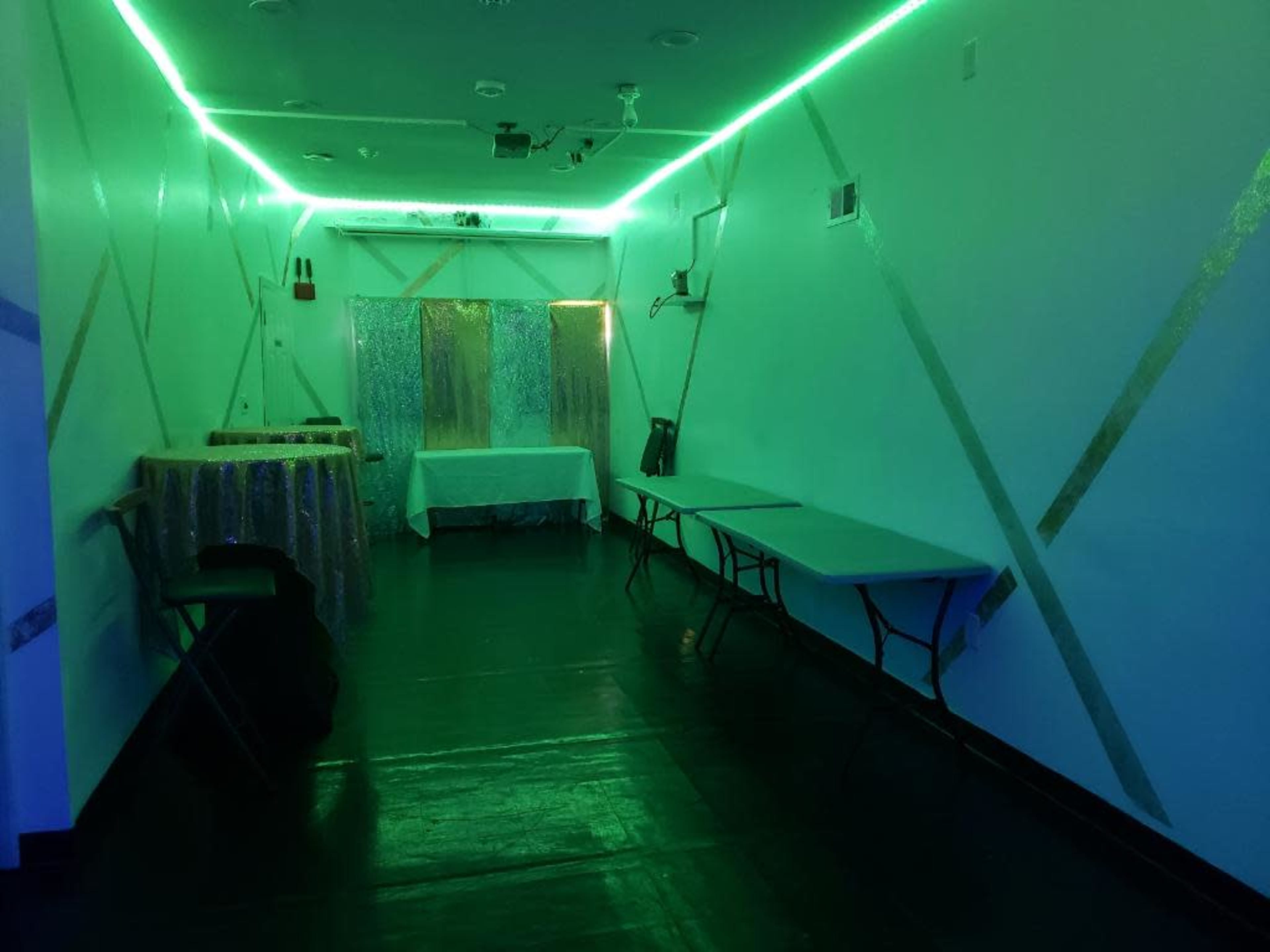 The image shows a dimly lit room with green neon lights, featuring two tables and a backdrop covered in shiny material.