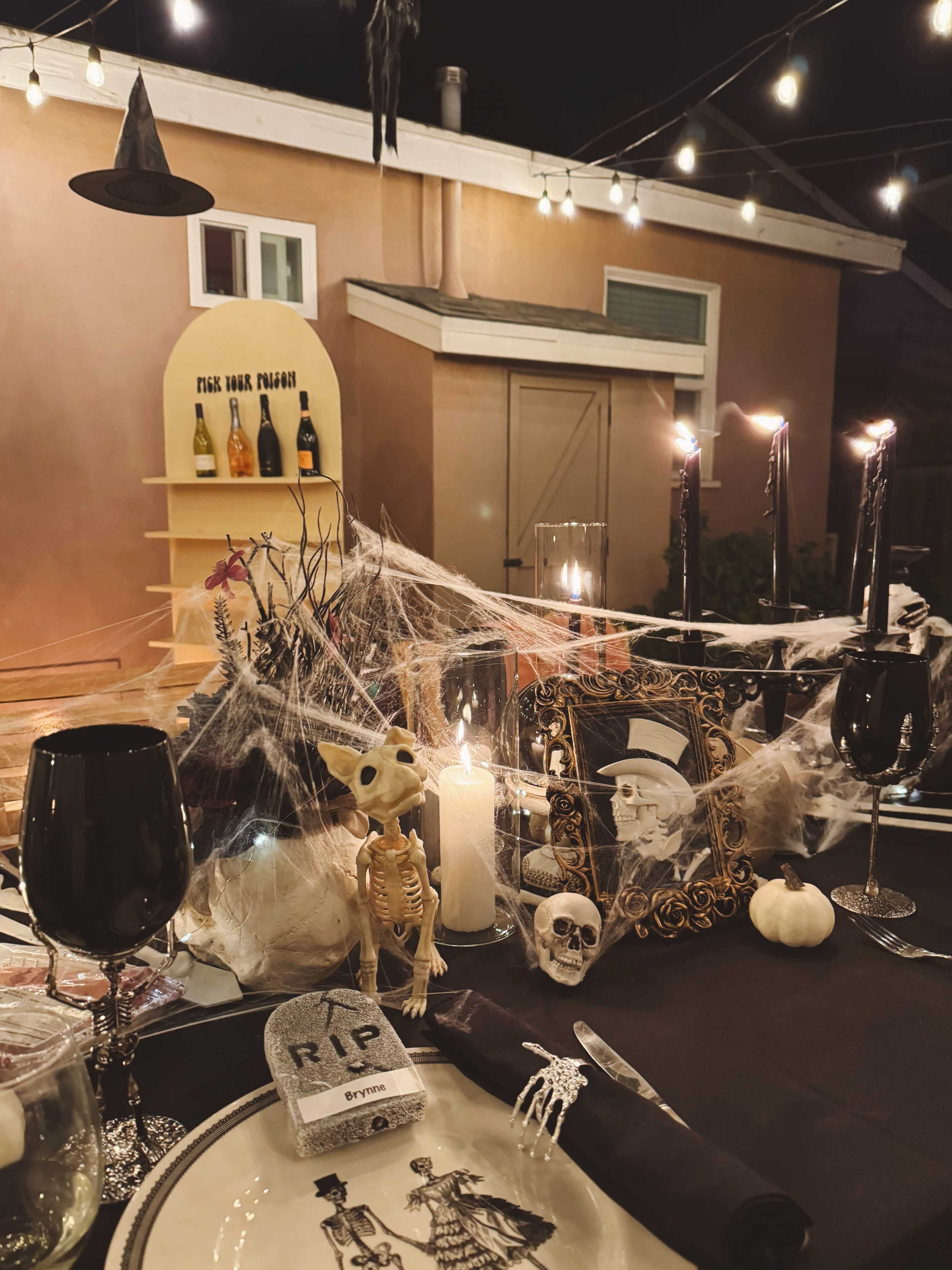 A Halloween-themed table is set with black plates, candles, skeleton decorations, and cobwebs, while a backdrop features bottles and string lights.