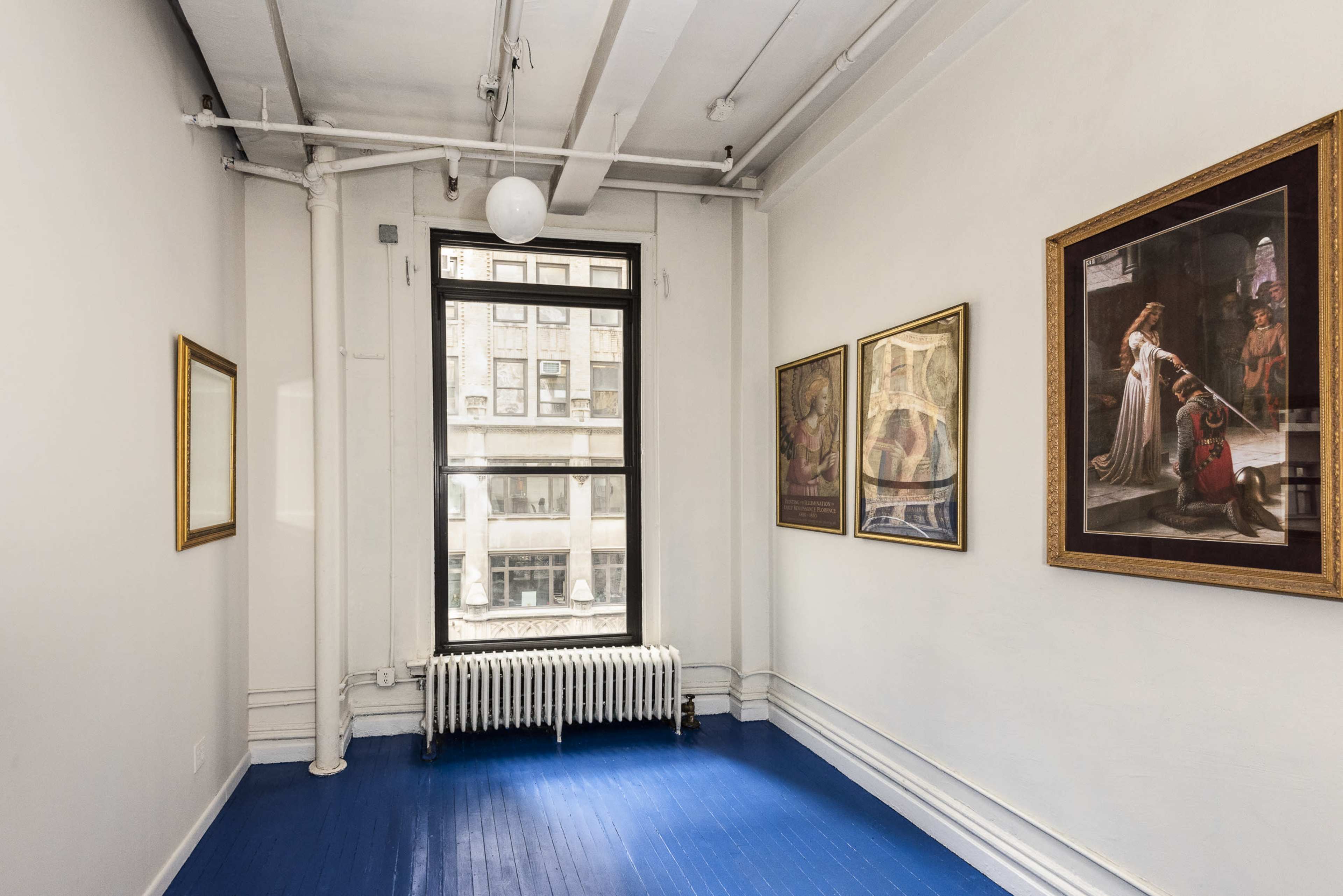 The image shows a small, brightly lit room featuring blue floorboards, two framed artworks on the walls, and a large window with a radiator beneath it.