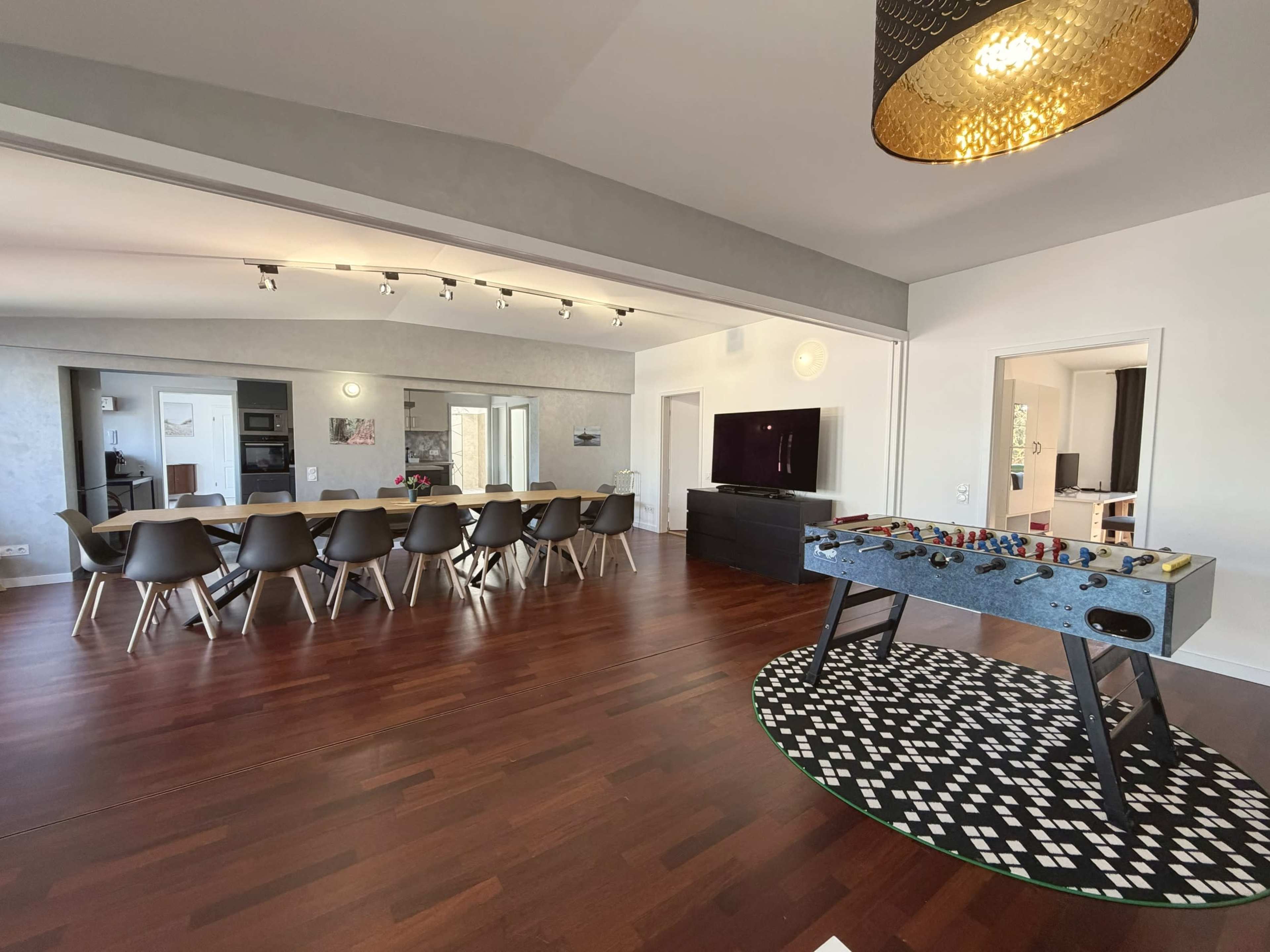 The image shows a modern open-concept living area featuring a dining table, a foosball table, and a television.