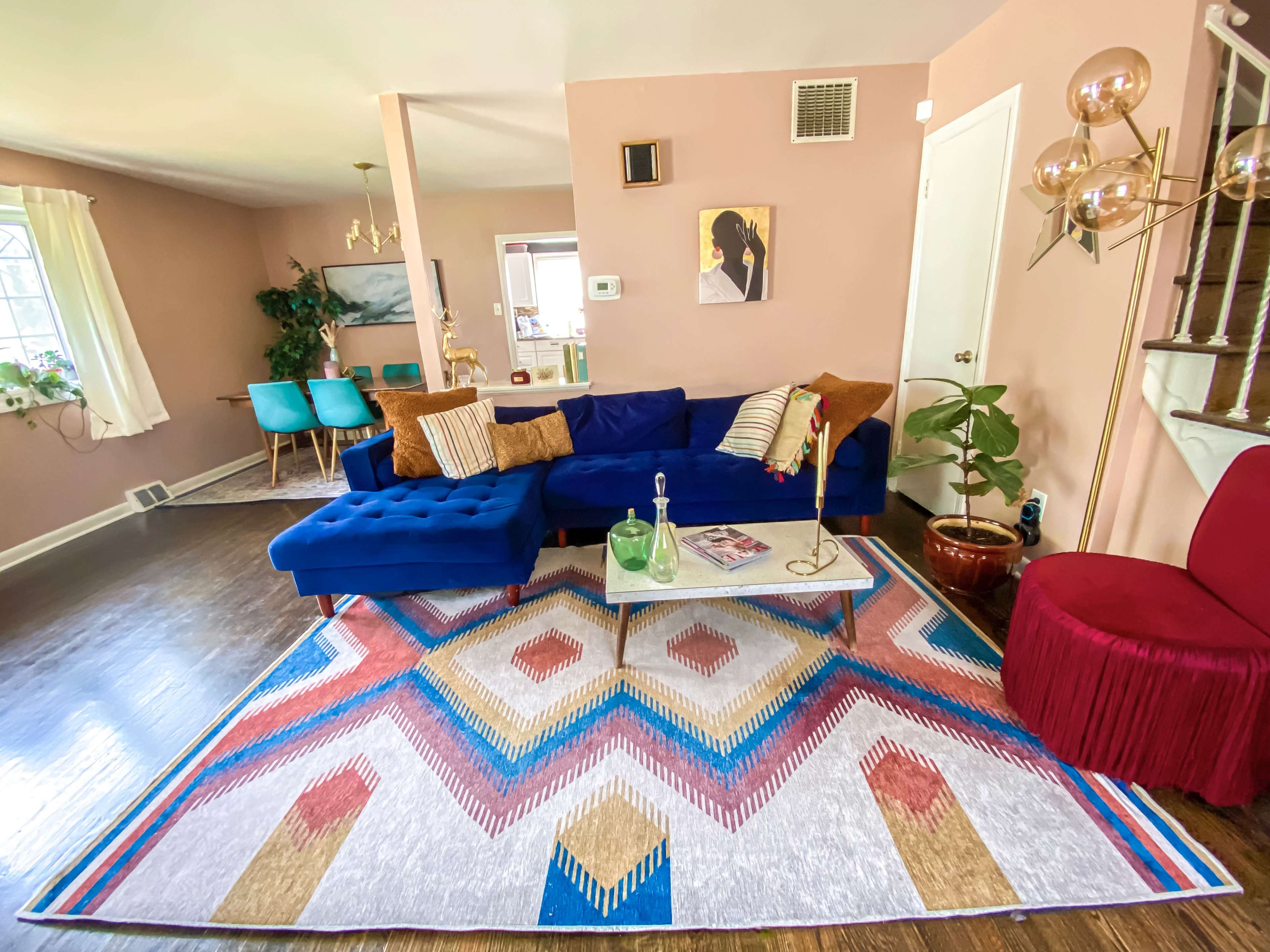 The image shows a bright living room featuring a blue sectional sofa, a geometric area rug, a glass coffee table, and a mix of decorative plants and furniture.