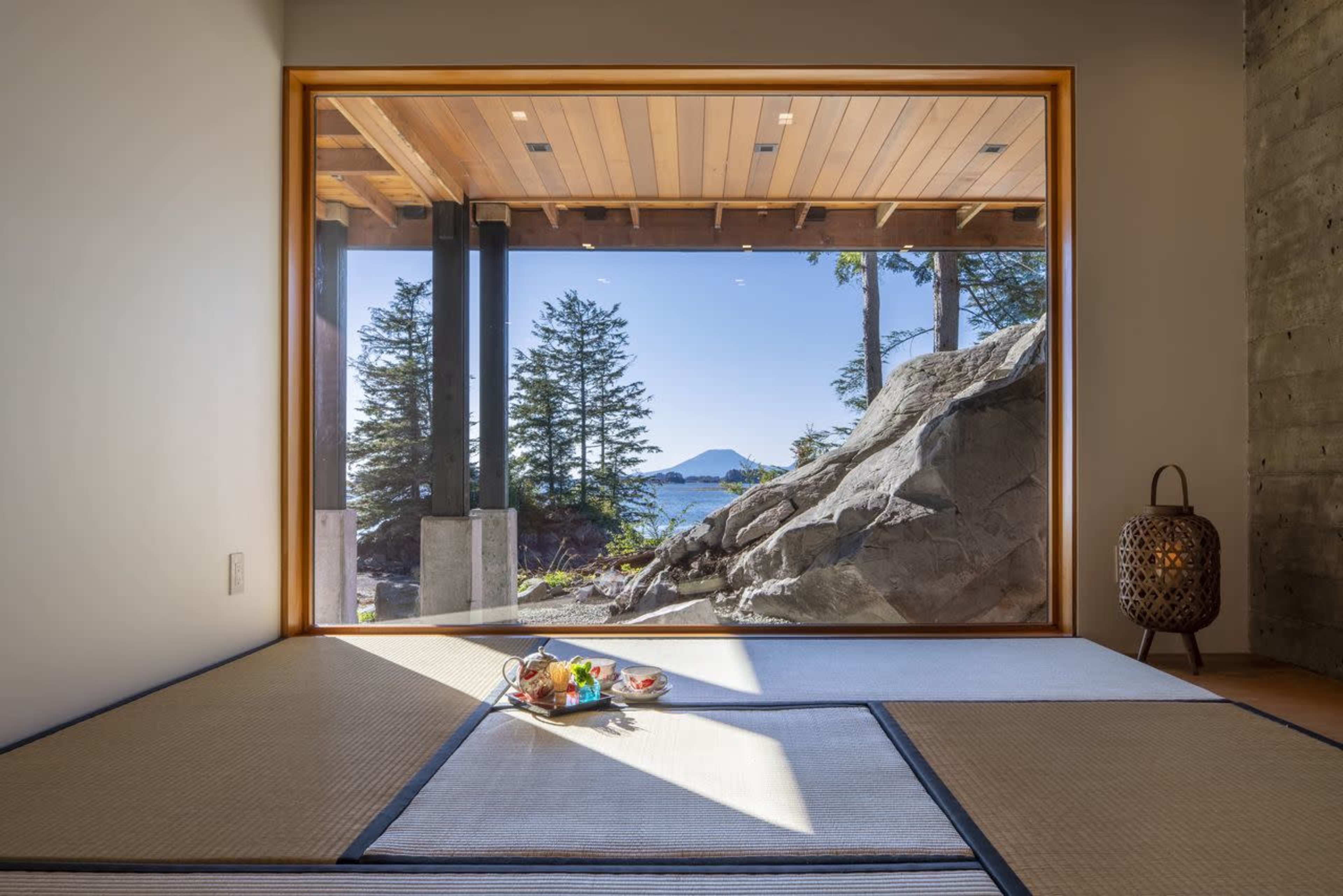 A spacious room features a large window overlooking a rocky landscape and distant mountain, with tatami mats and a decorative arrangement in the foreground.