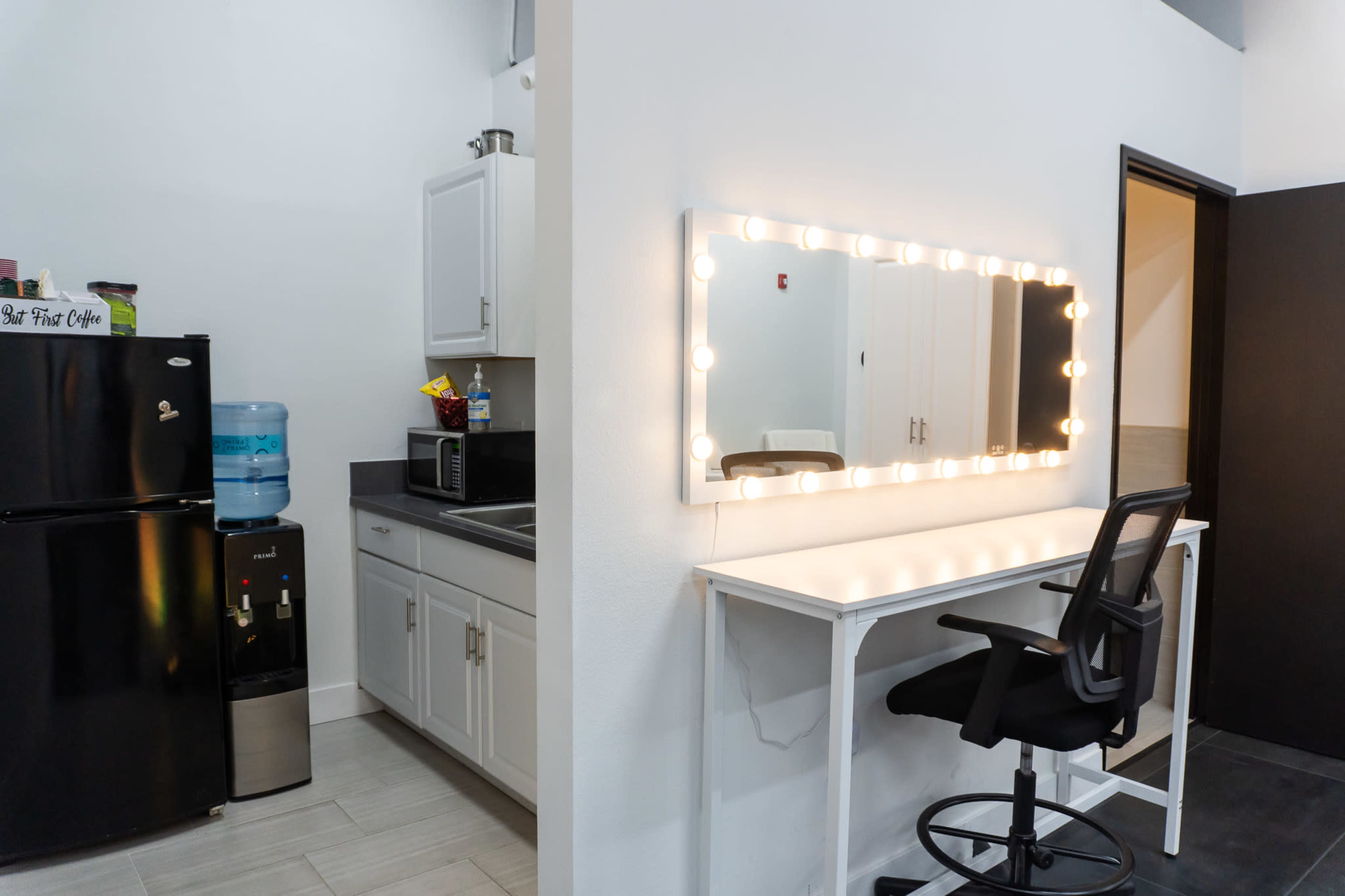The image shows a kitchenette area with a refrigerator and microwave on one side, and a simple vanity with a large illuminated mirror and a chair on the opposite wall.