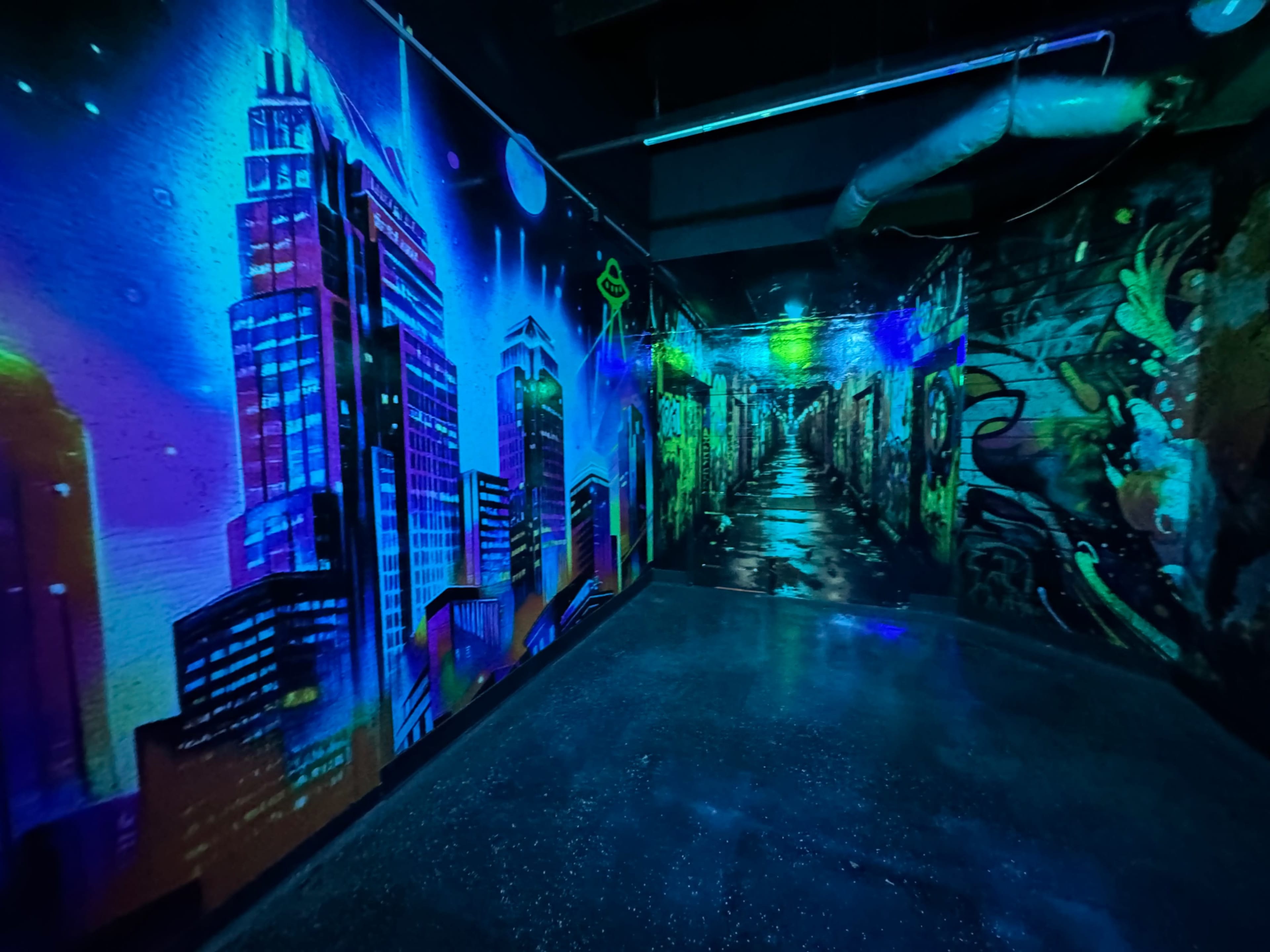 The image depicts a brightly colored mural featuring urban landscapes and graffiti, illuminated by neon lights in a dimly lit corridor.