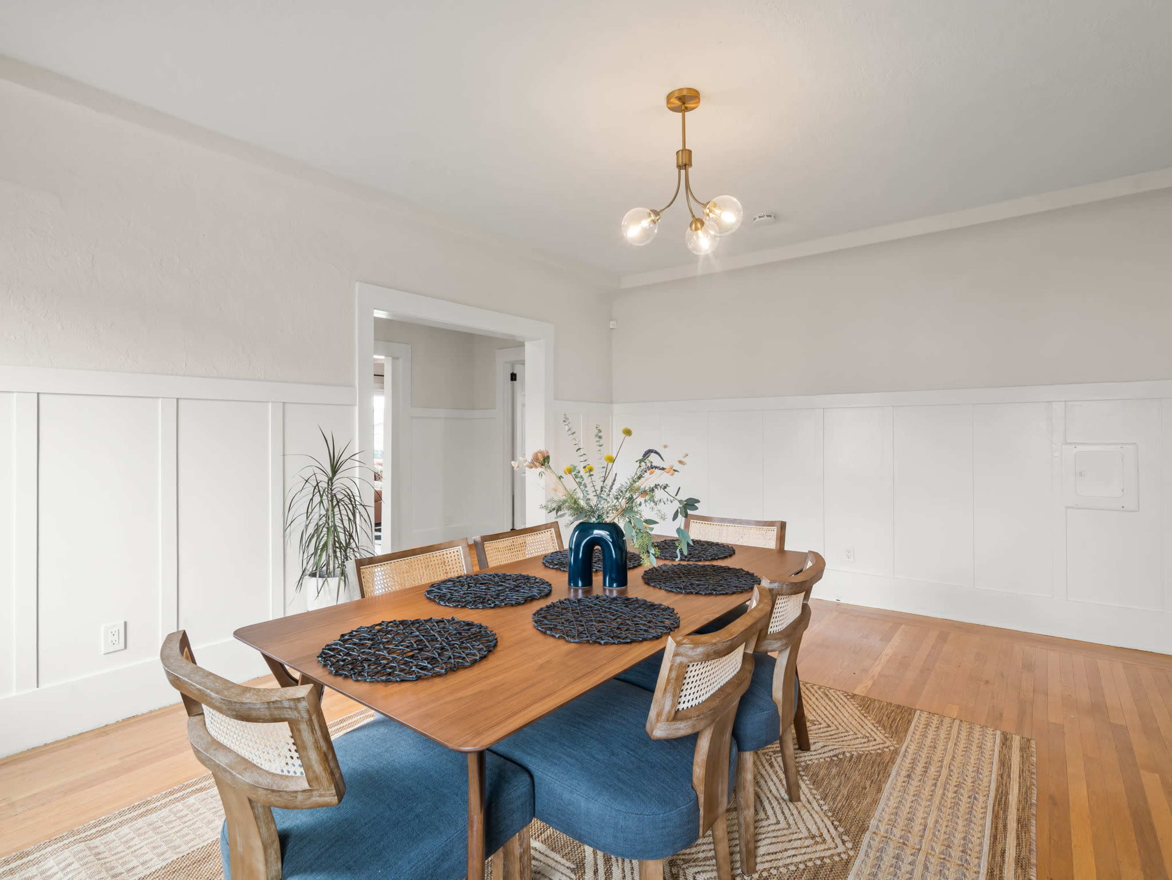 A spacious dining room features a wooden table surrounded by eight chairs, with decorative placemats and a centerpiece of flowers under a modern light fixture.
