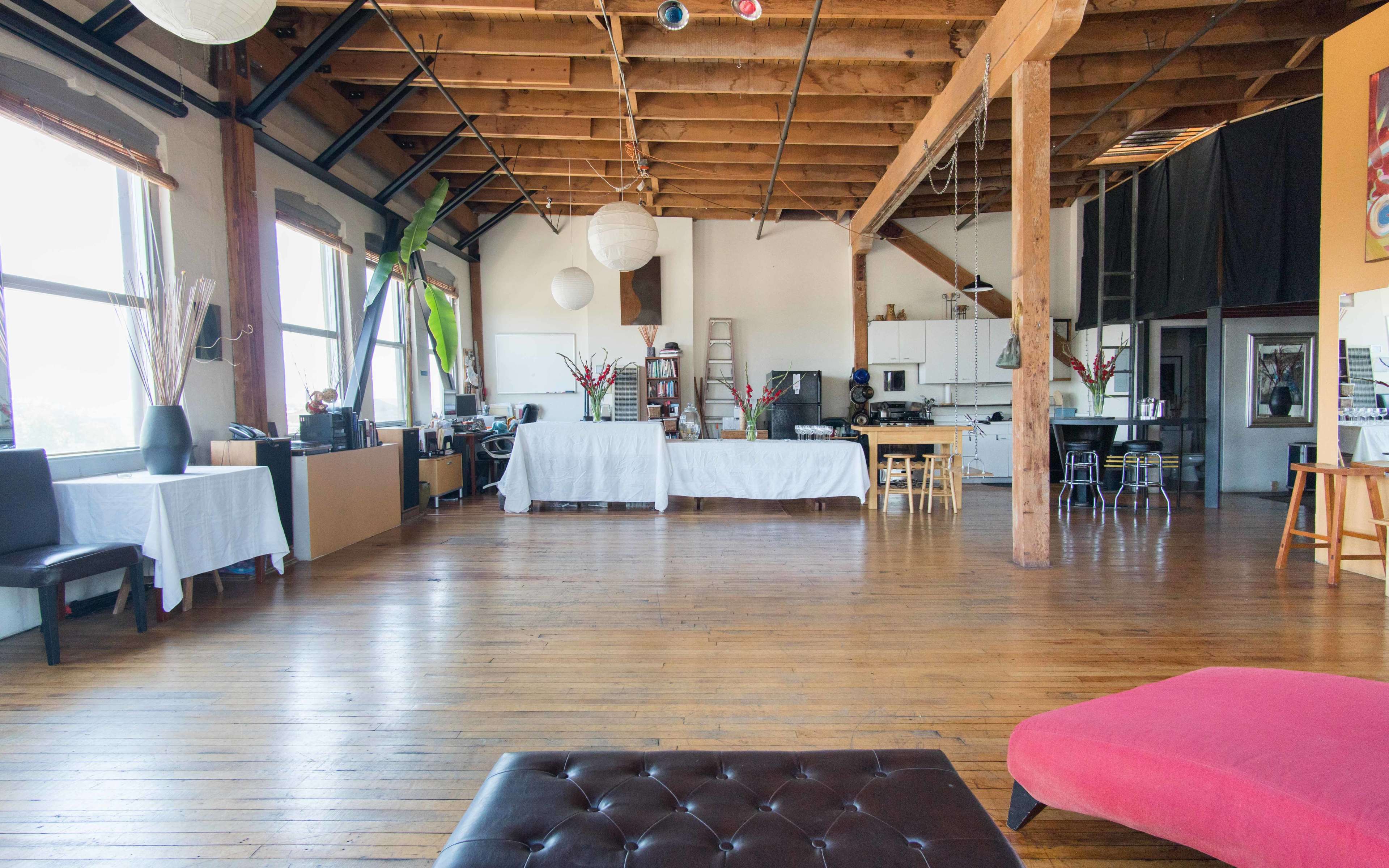 A spacious, open loft with wooden floors, exposed beams, and large windows, featuring a kitchen area and tables decorated with white tablecloths.