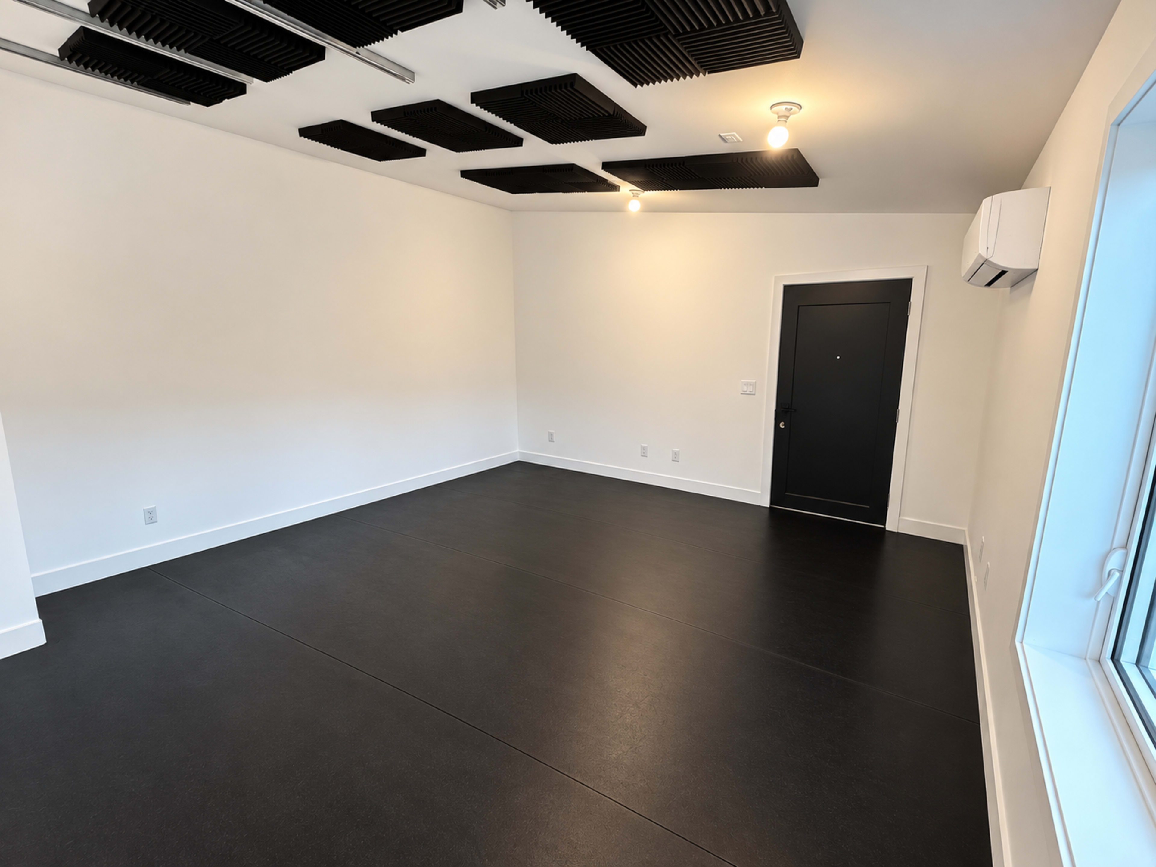 The image shows a sparsely furnished room with black flooring, white walls, and soundproofing panels on the ceiling.