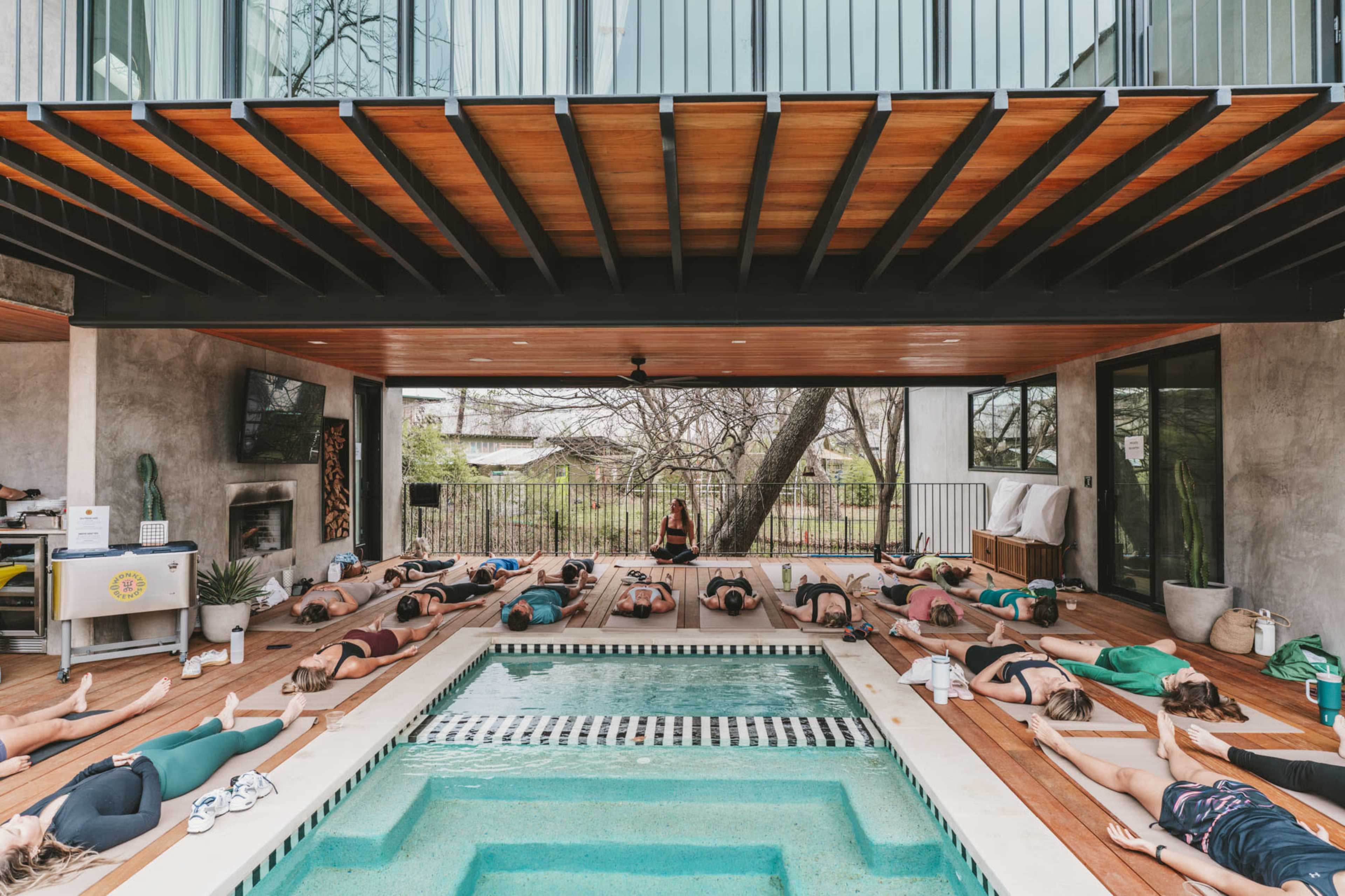 A group of individuals is practicing yoga on mats placed around a pool in a spacious, modern indoor-outdoor setting.