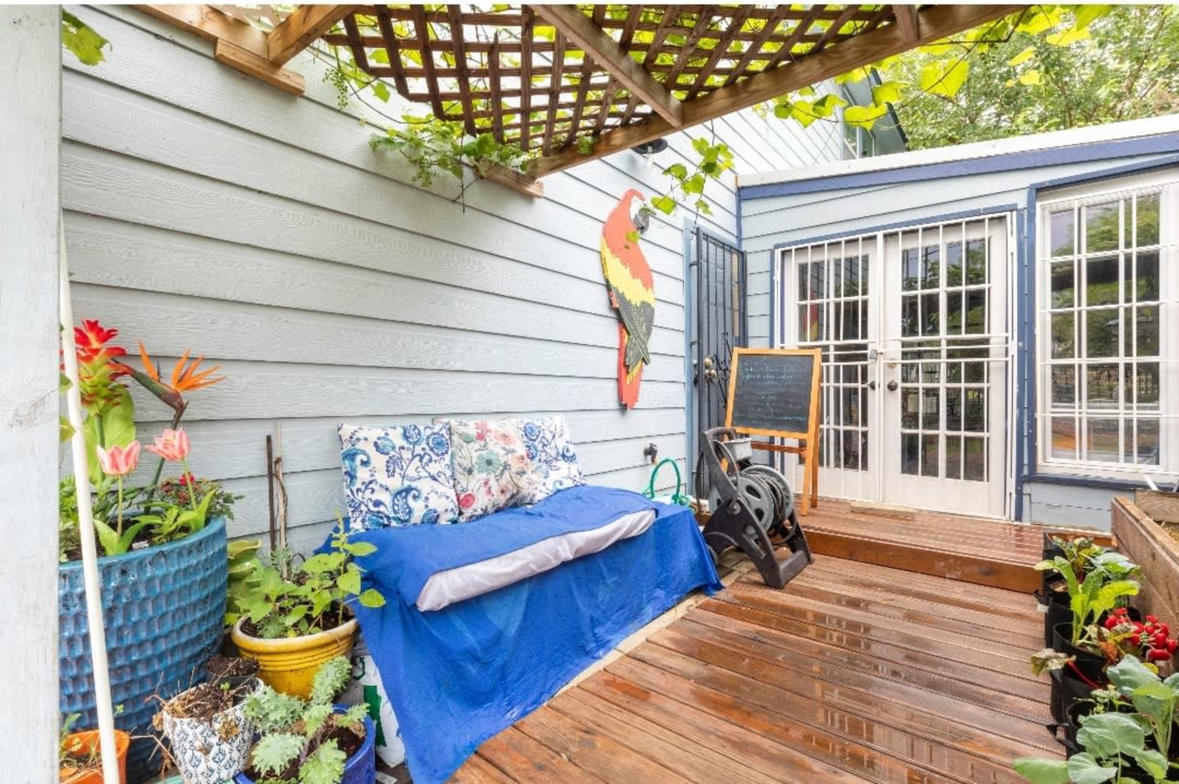 A cozy outdoor seating area with a wooden deck, surrounded by potted plants and a partially covered space with a blue blanket draped over a bench.