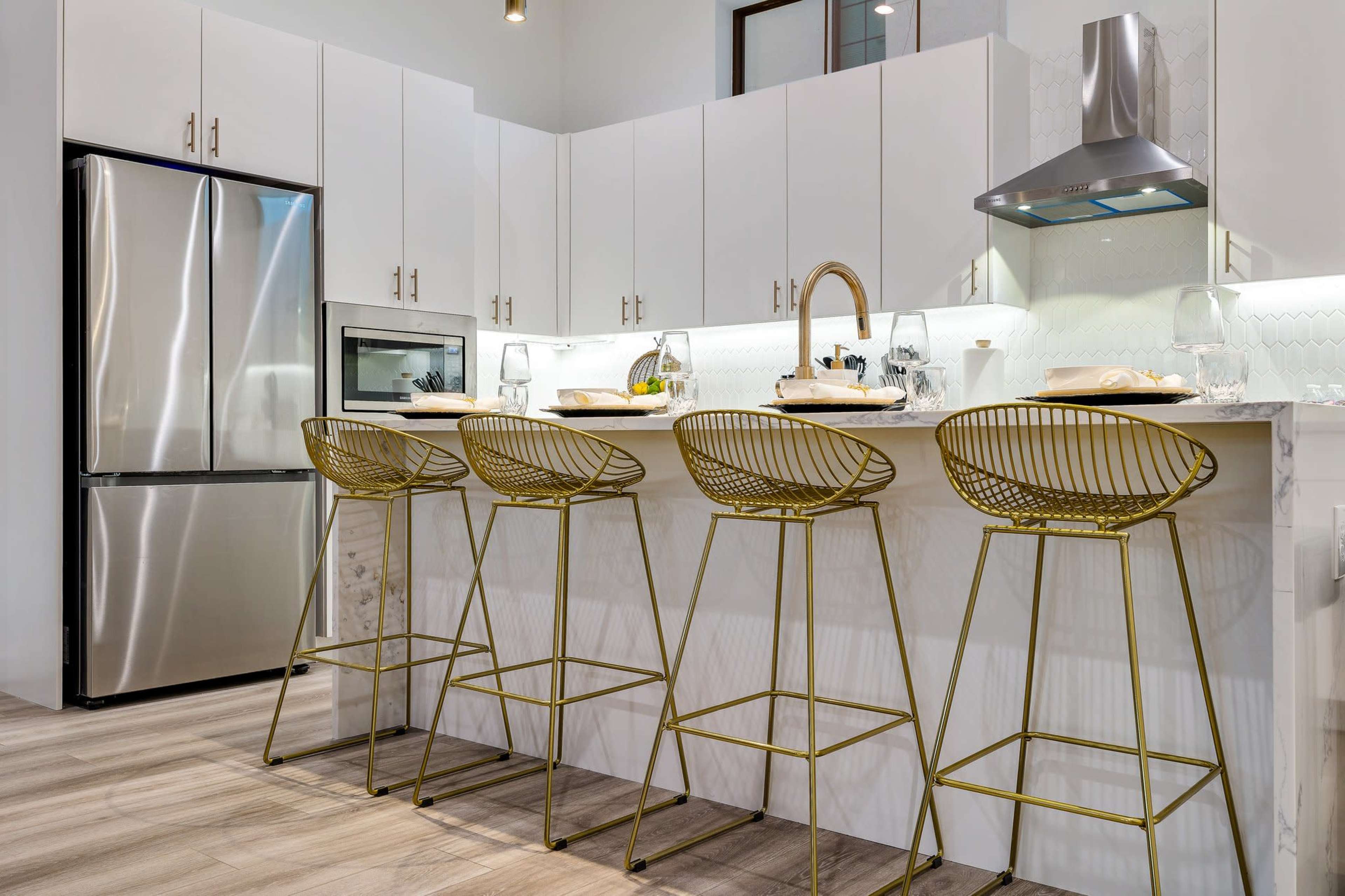 A modern kitchen features a marble countertop with four gold bar stools and stainless steel appliances.
